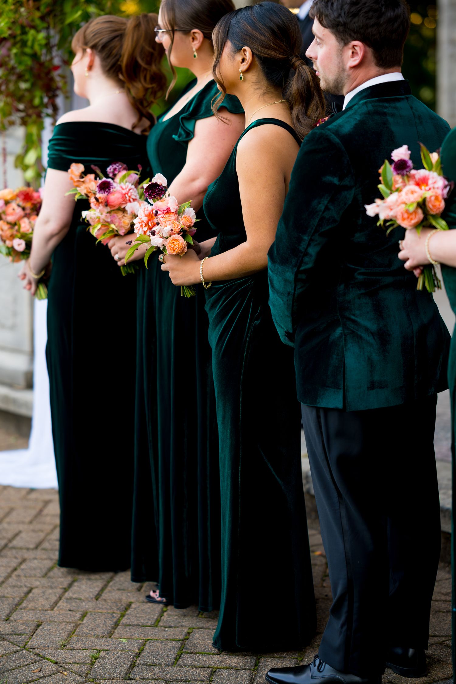Wedding party members in formal black attire stand in line holding colorful coral and pink floral bouquets.