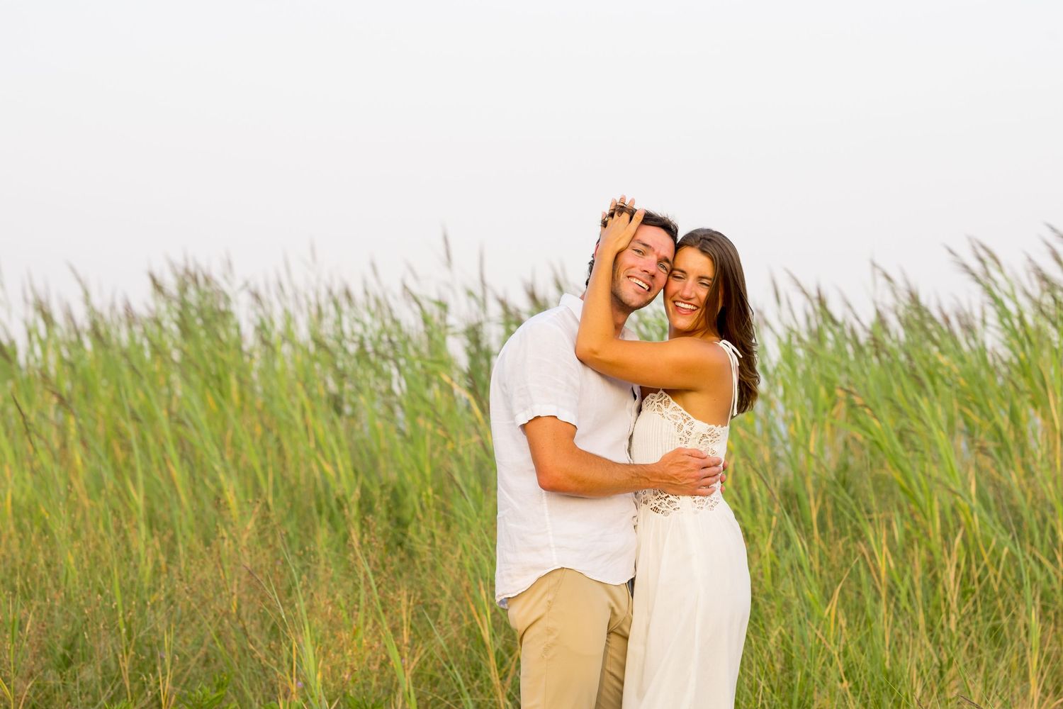 Couple embraces while standing in tall grass field during golden hour.