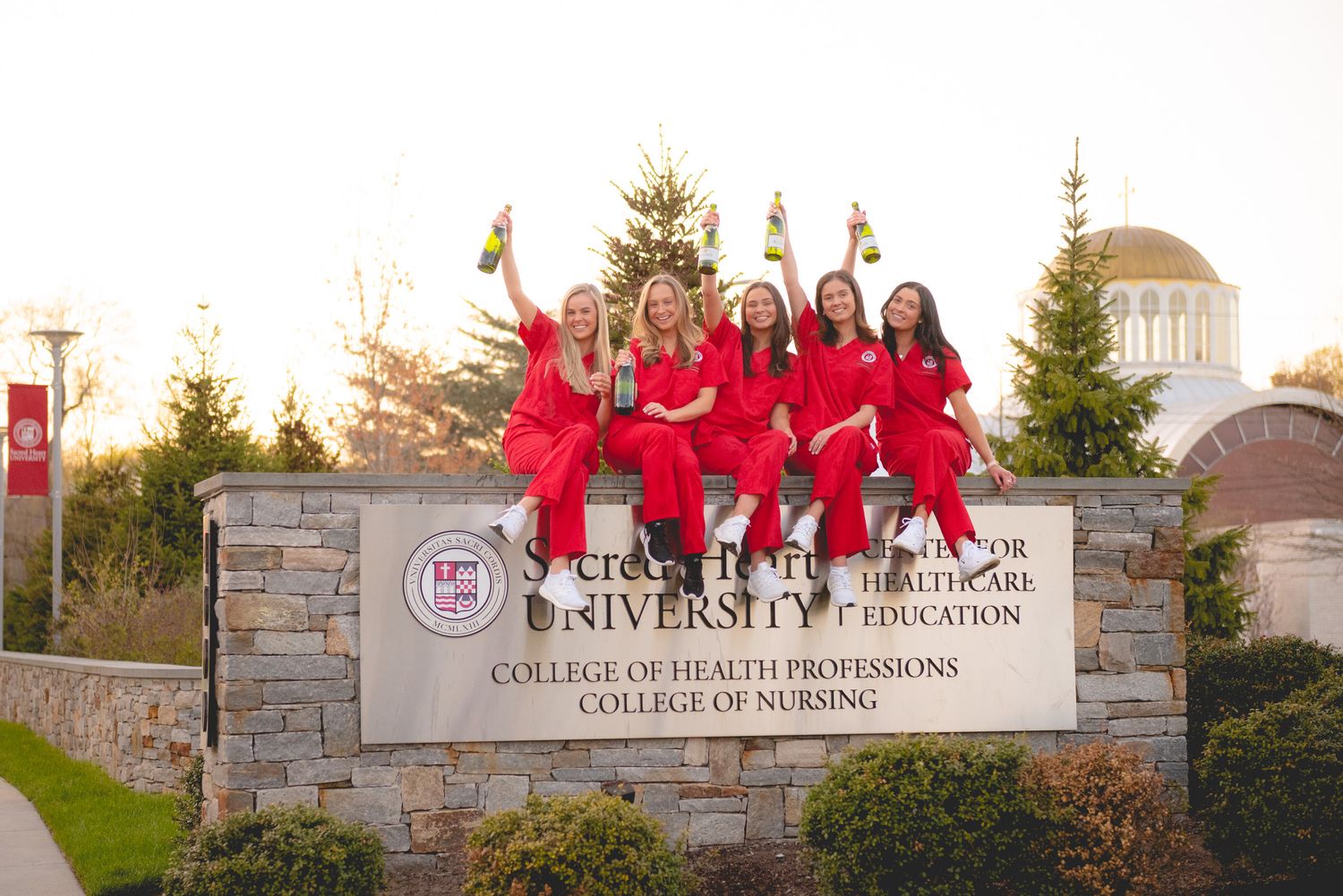 Sacred Heart nursing graduates in red scrubs celebrate on university sign wall at sunset for graduation photos.