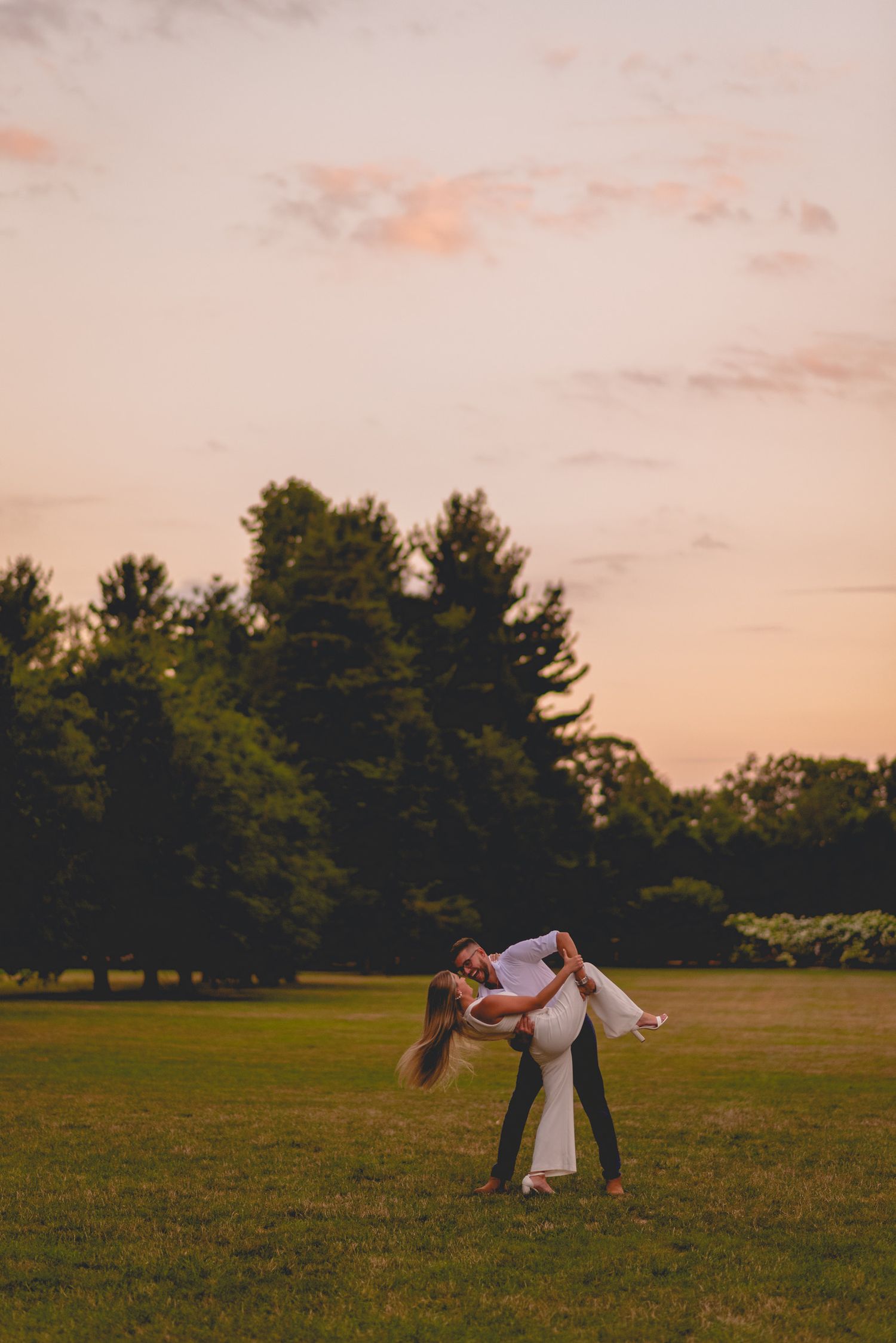 A couple dances playfully in an open field at sunset with trees in the background.