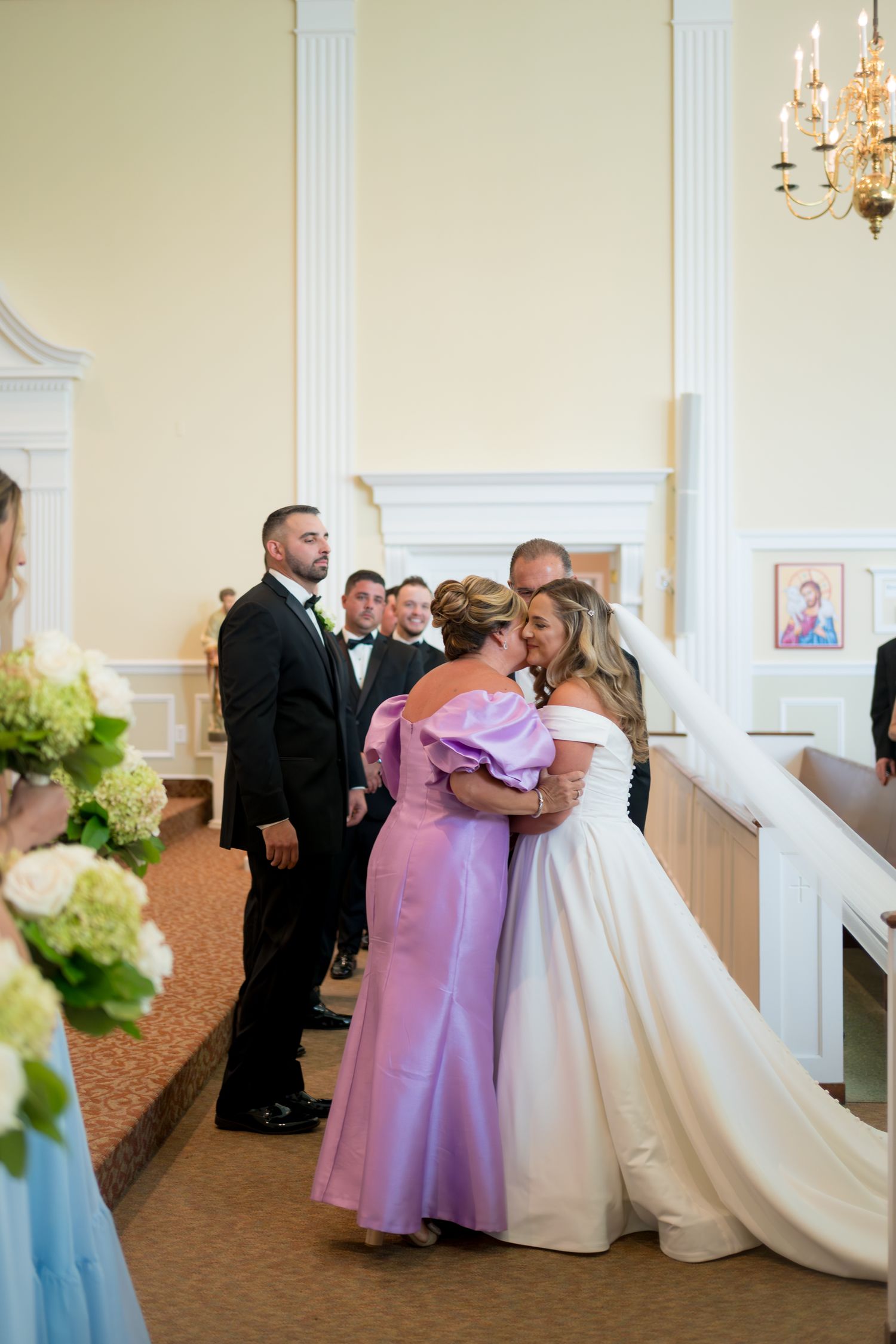 Wedding party members in formal attire gather in an elegant room with white columns and chandeliers.