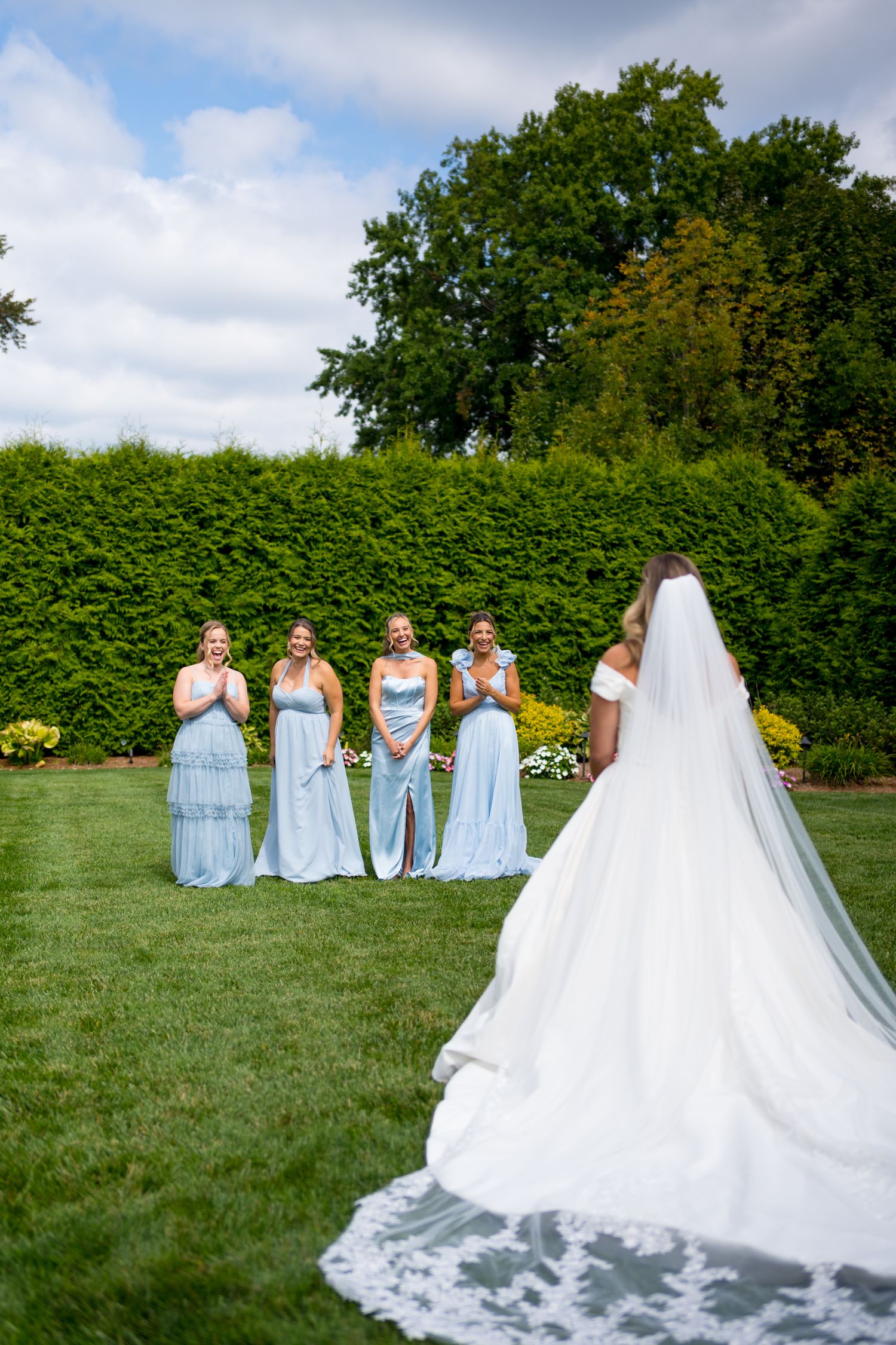 A wedding party in light blue bridesmaid dresses poses on a grassy lawn with trees in the background.