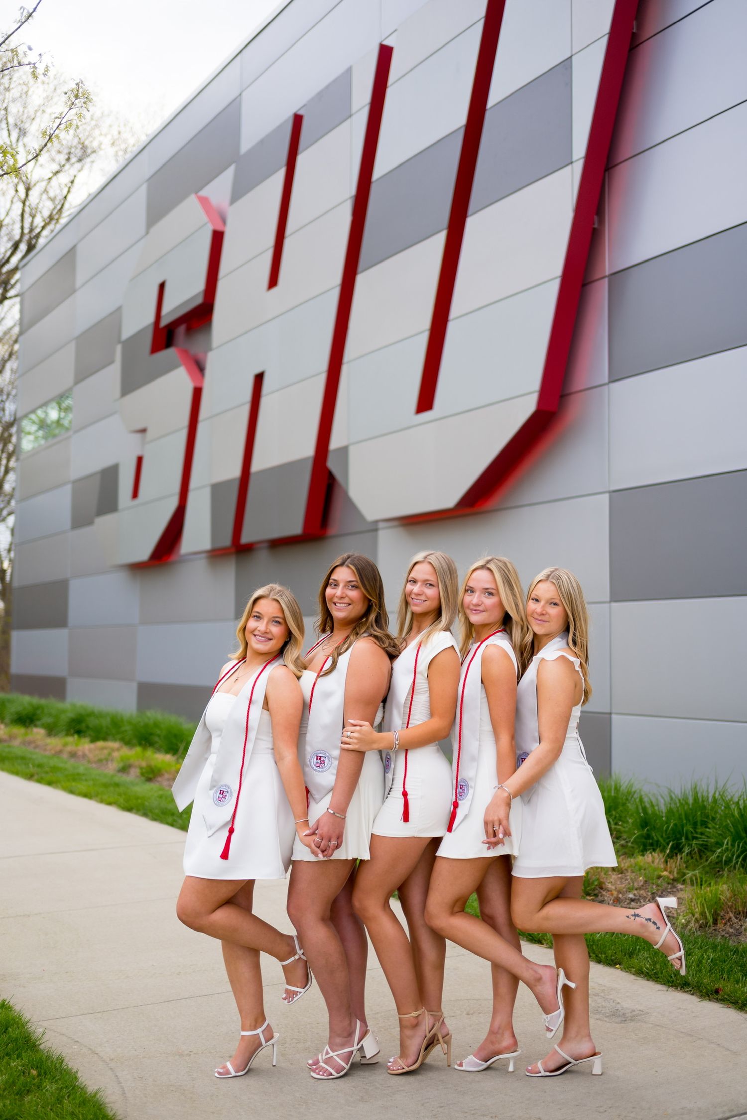 Sacred Heart University graduates in white outfits against a modern geometric wall with red accents.