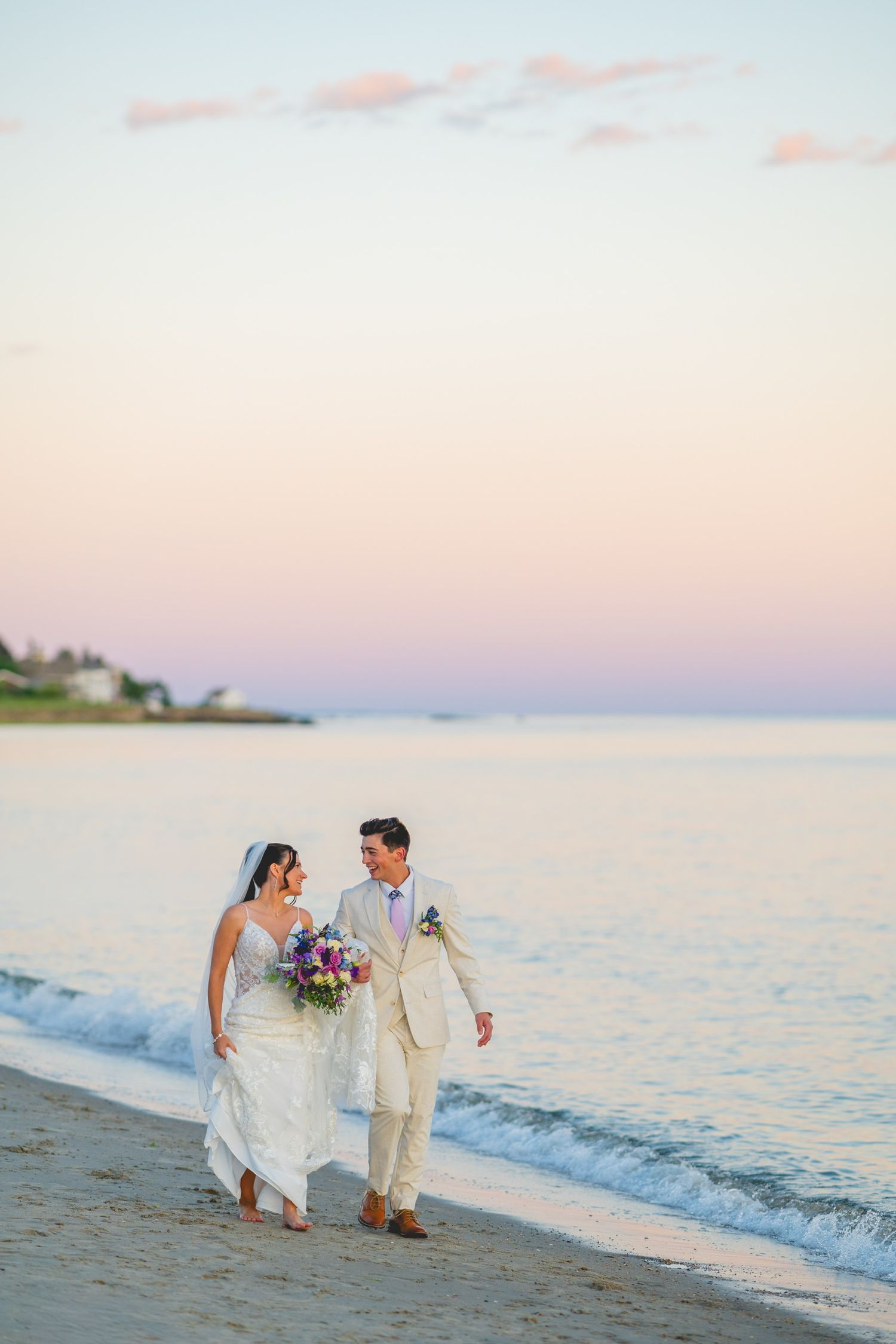 Couple walking along a scenic beach at sunset in wedding attire with ocean waves in the background.