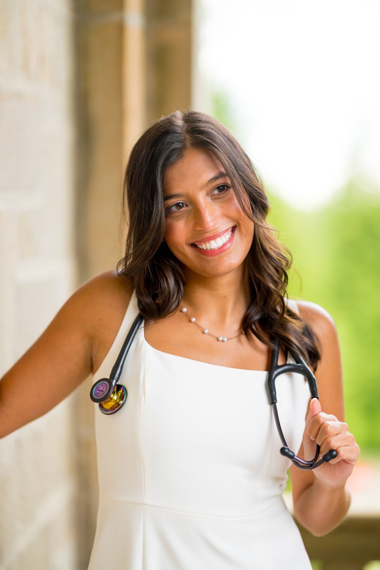 Fairfield University graduate in white attire holding stethoscope while posing against outdoor background.