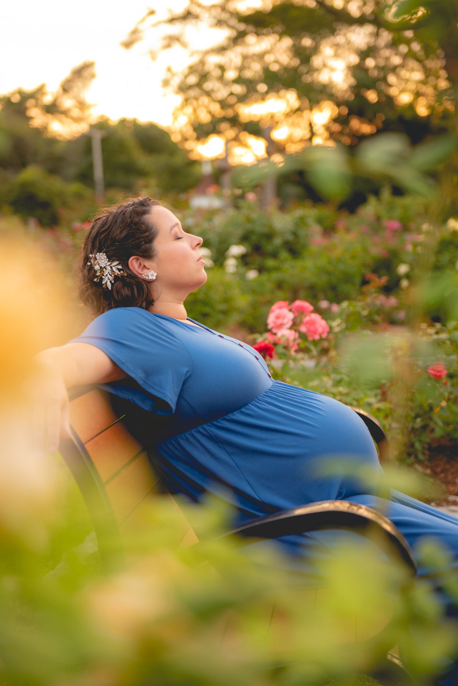 Pregnant woman in blue dress relaxing in a rose garden during golden hour.