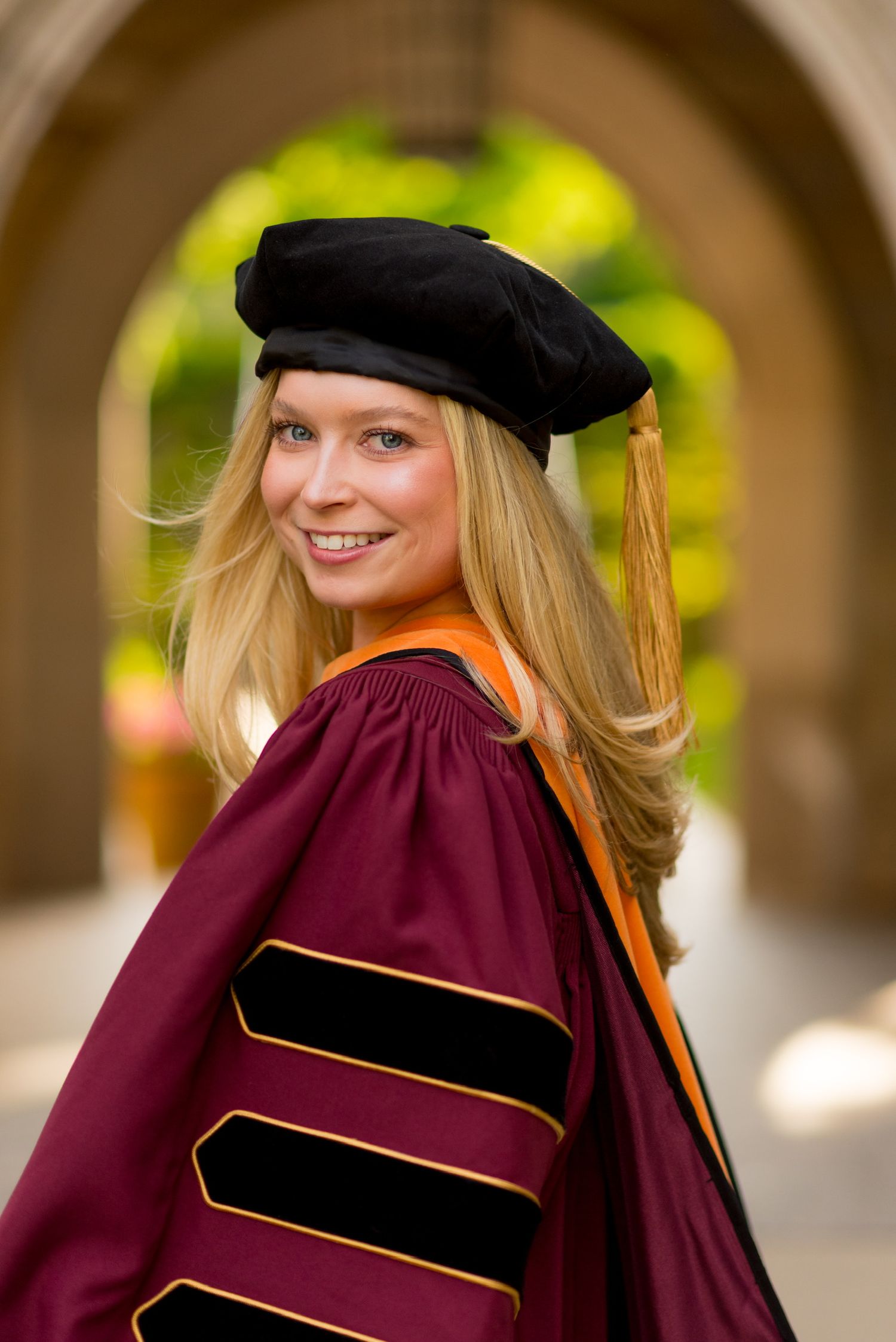 Graduate in burgundy doctoral regalia and cap poses under stone archway.