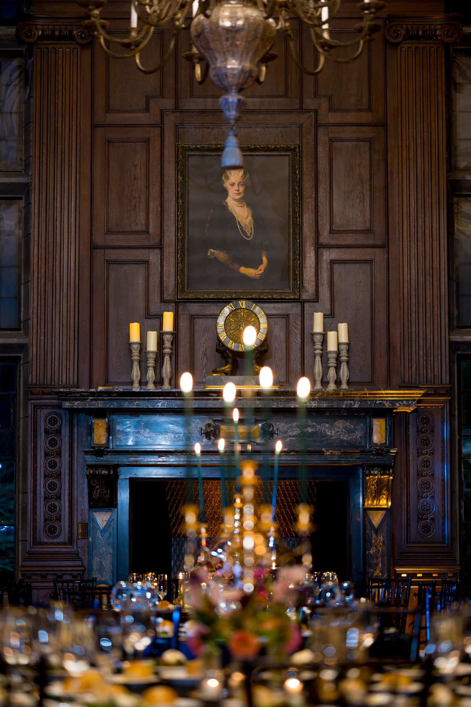 Elegant dining room with wood paneling, portrait above fireplace, and candlelit table settings.