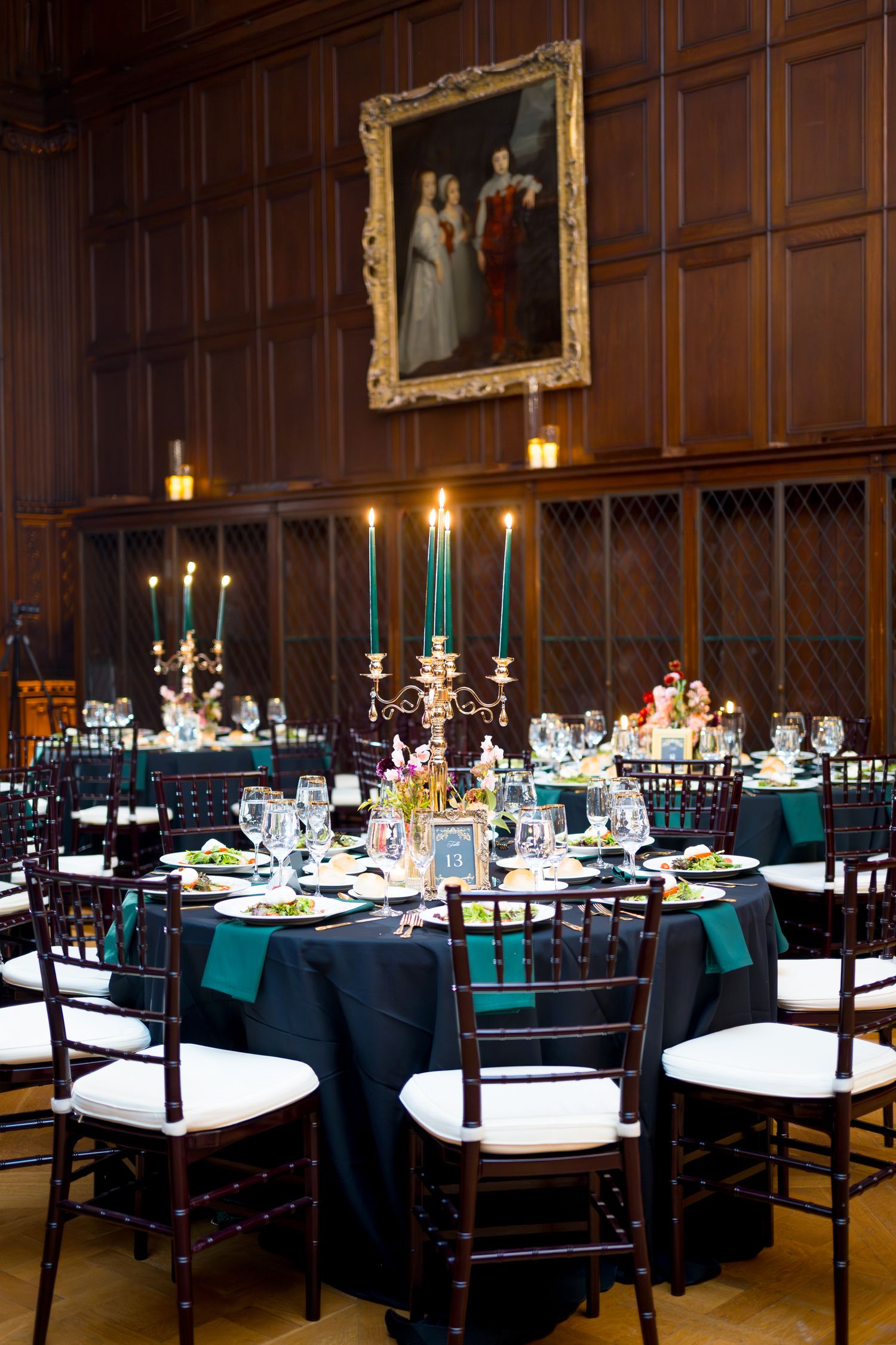 Elegant dining tables set with black chairs, teal linens, and glowing candelabras in a wood-paneled banquet hall.