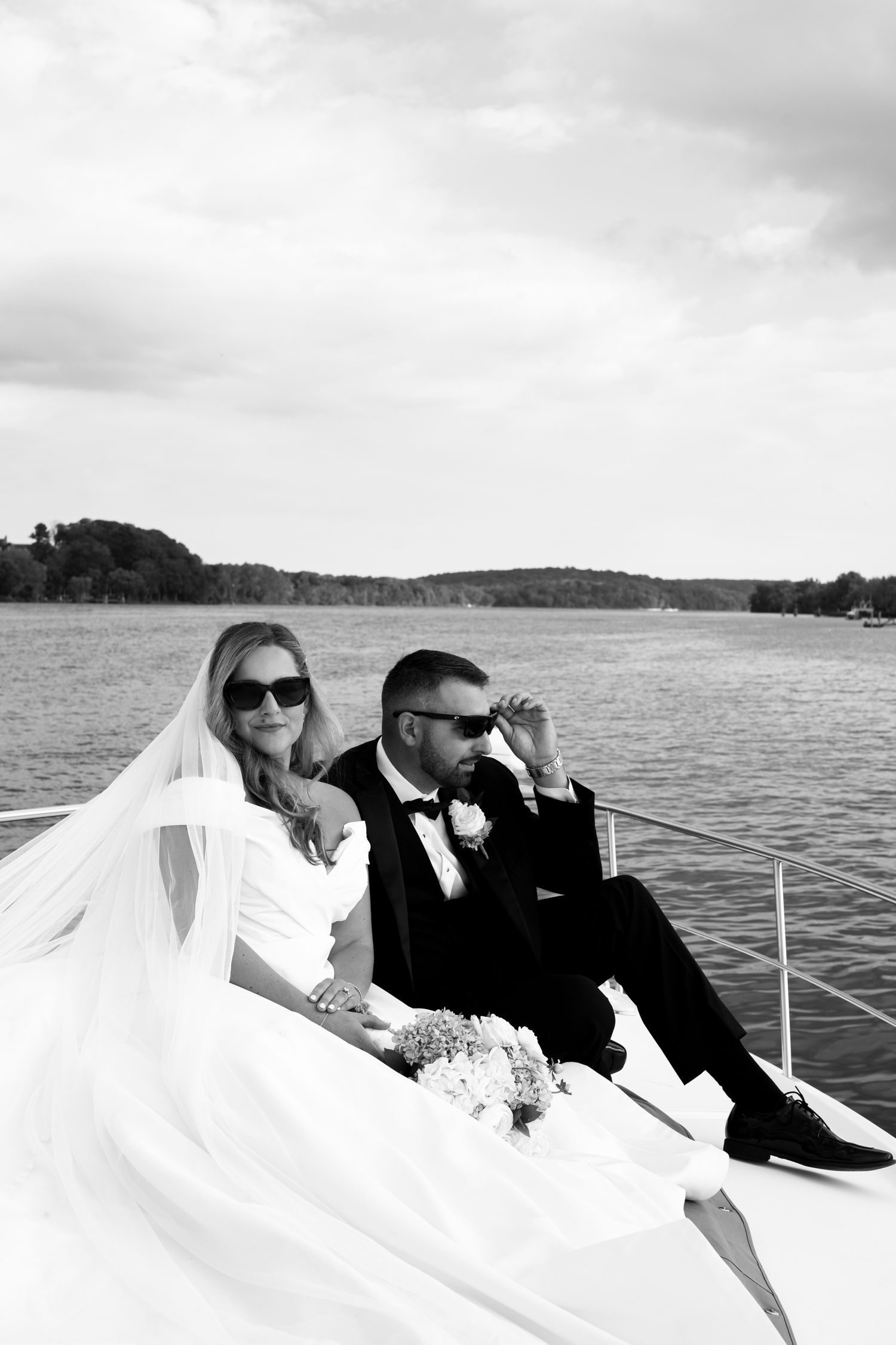 Newlywed couple in formal attire relaxing on boat deck with waterfront view.
