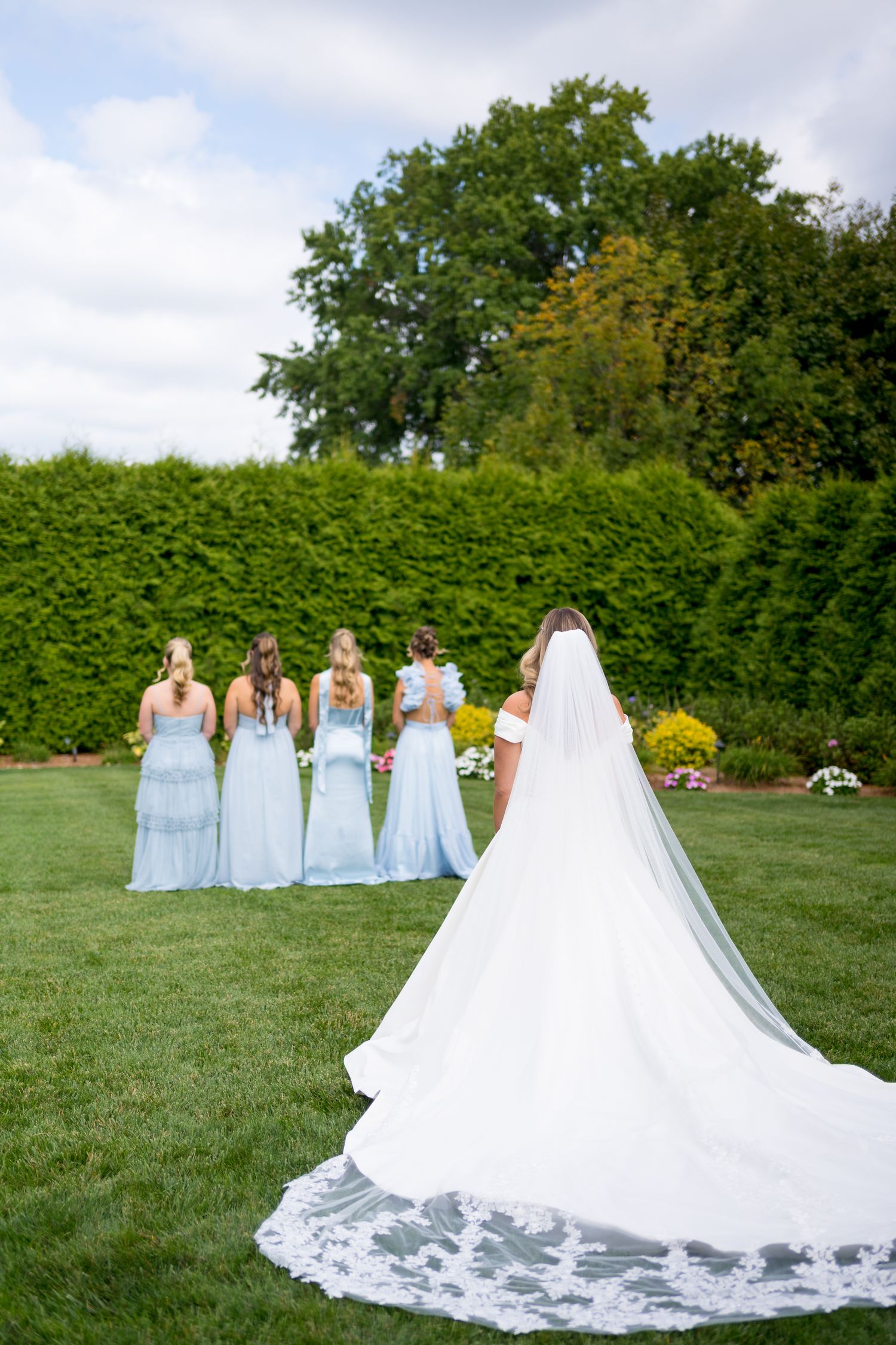 A bride in a flowing white gown with long train stands with bridesmaids in pale blue dresses on a green lawn.