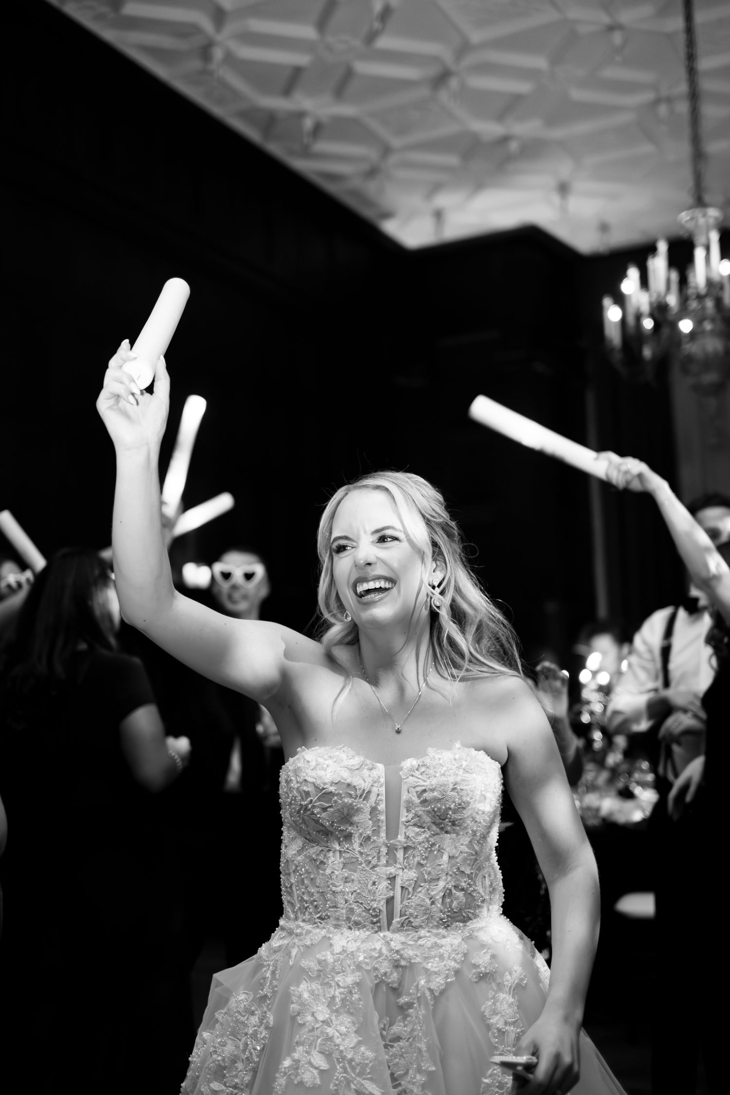 A bride in a white lace dress dances joyfully at her wedding reception under ornate chandeliers.