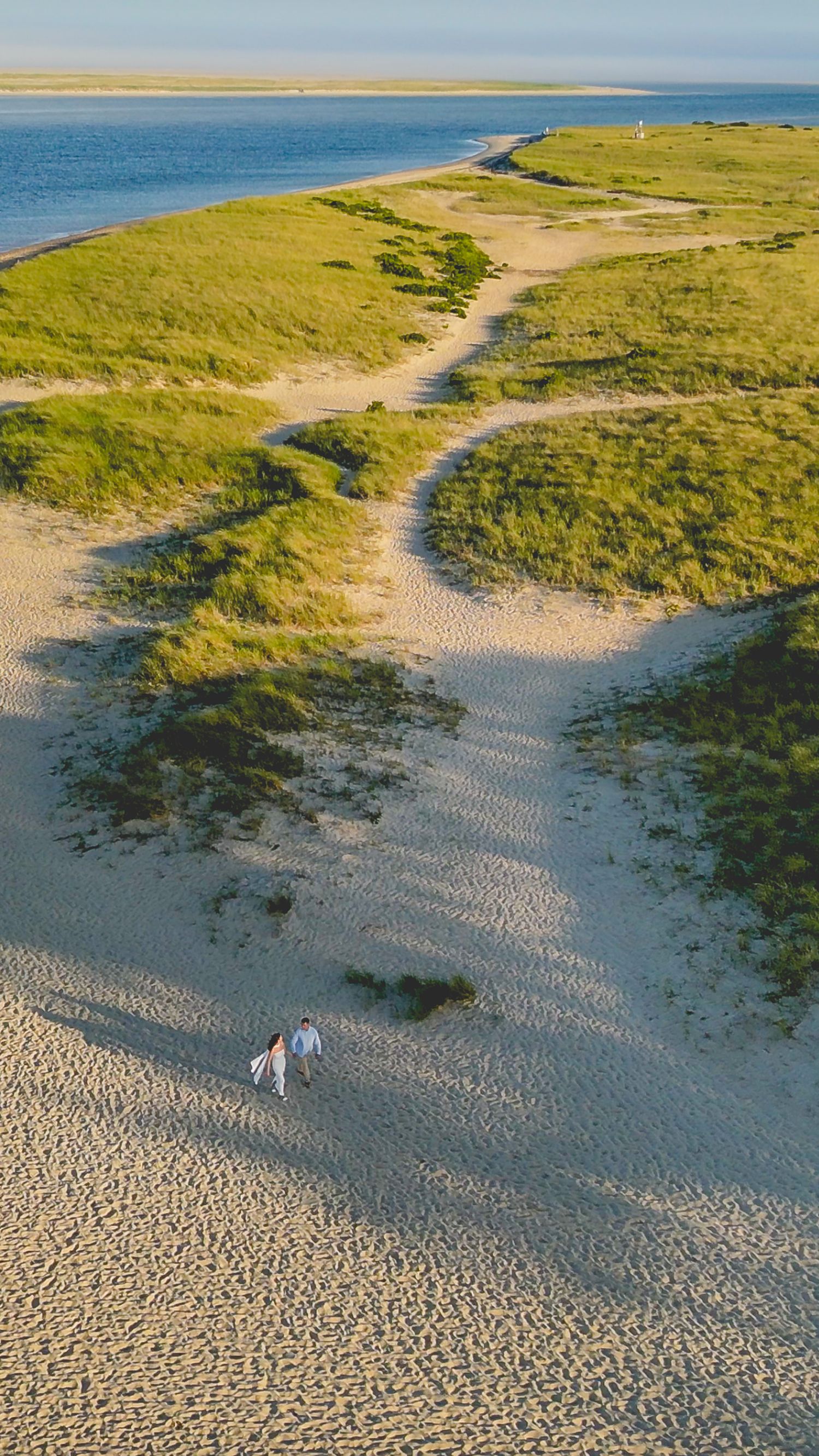 Aerial view of sandy beach path winding through coastal dunes with two people walking near the ocean.