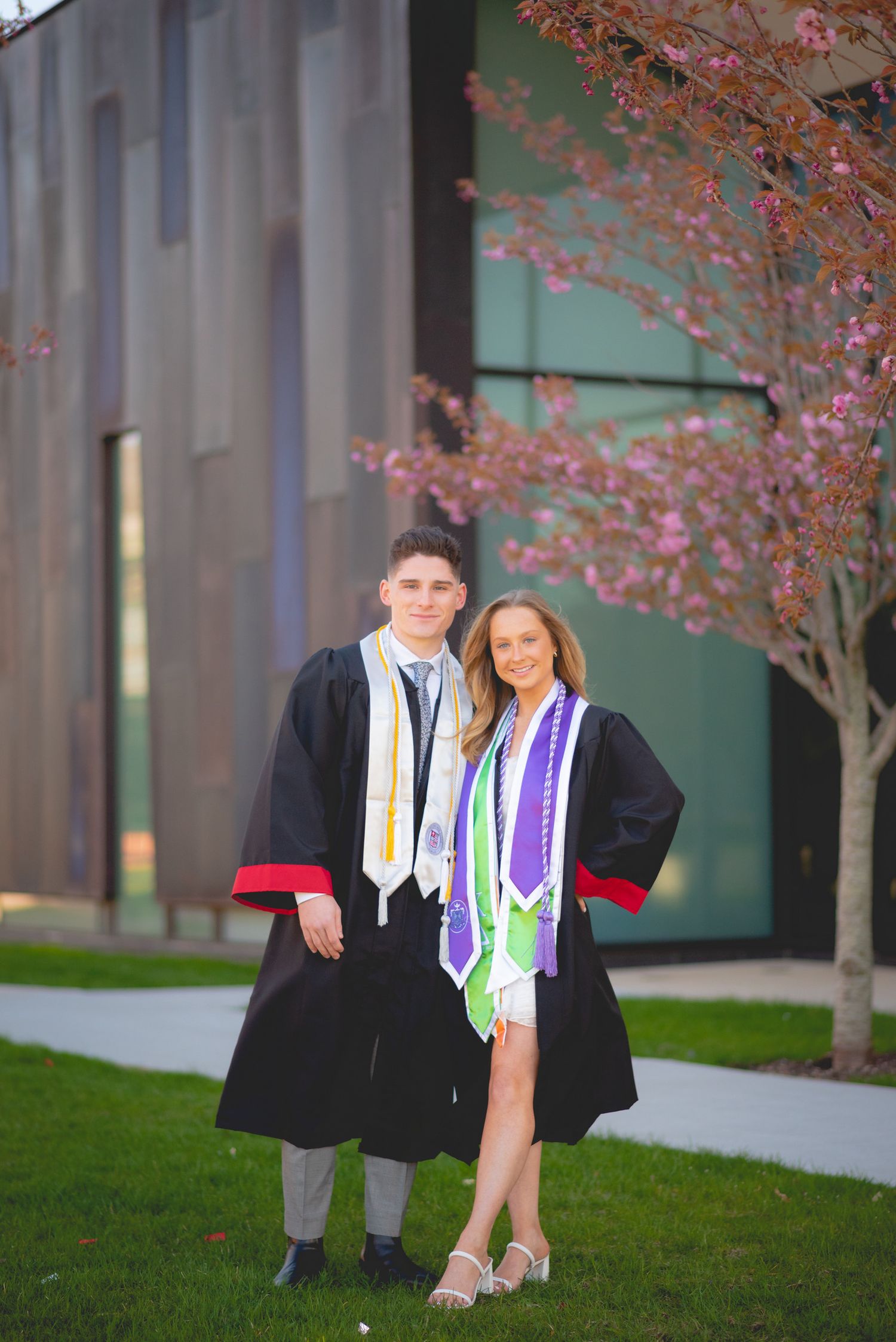 Two Sacred Heart University graduates in black robes and colorful stoles pose together under blooming cherry blossom trees.