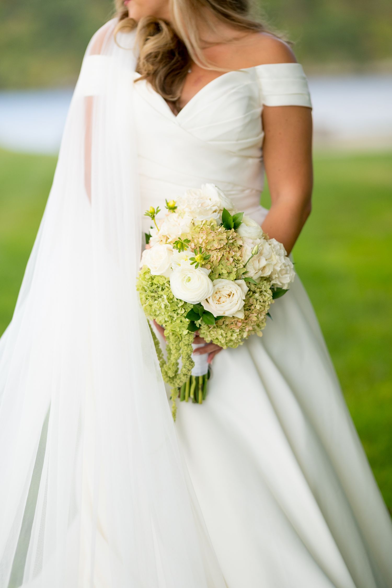 Close-up of bride's white off-shoulder wedding dress and bouquet featuring white roses and green hydrangeas.