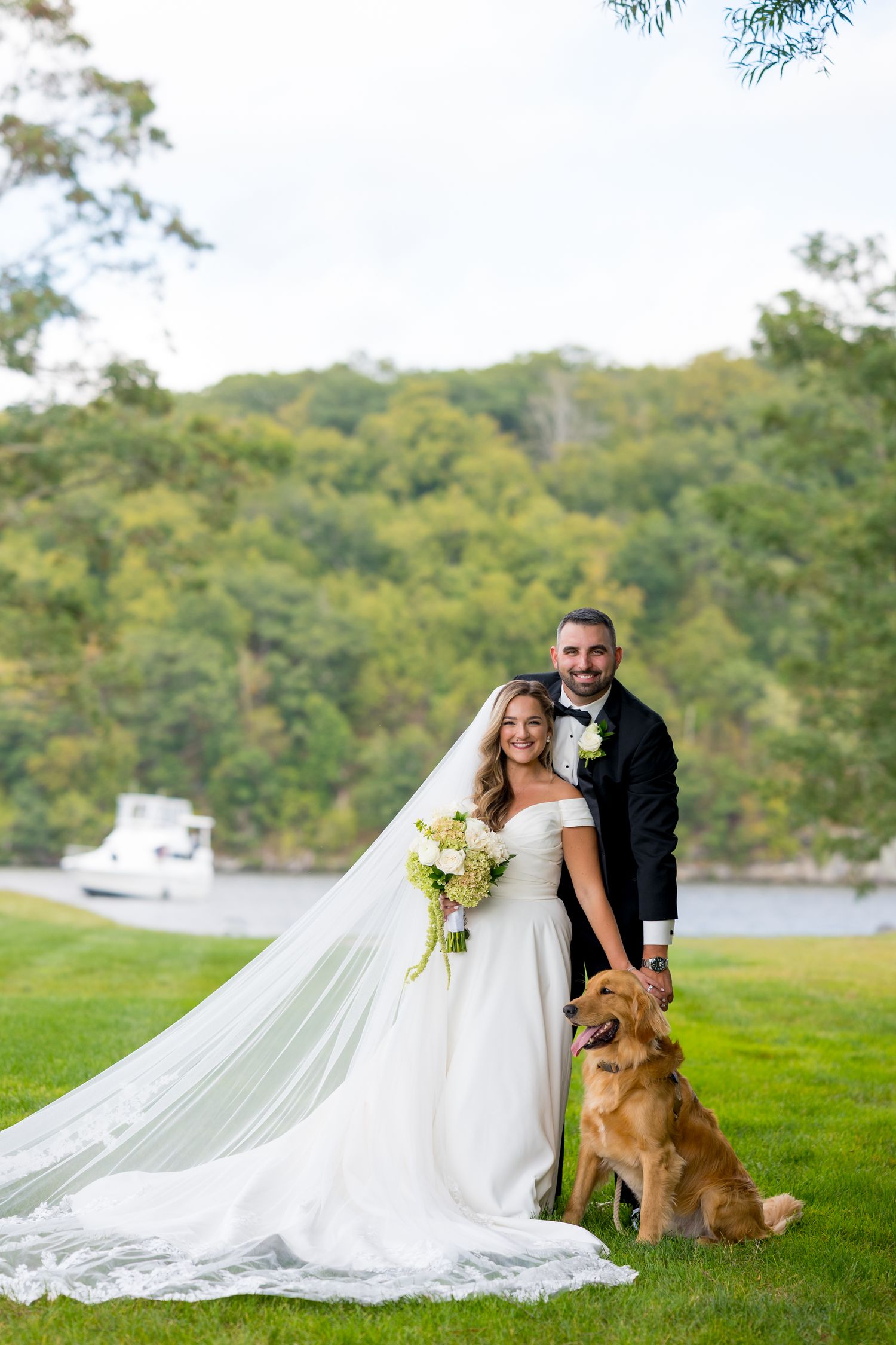 Wedding couple posing with their golden retriever on grassy lawn near waterfront.