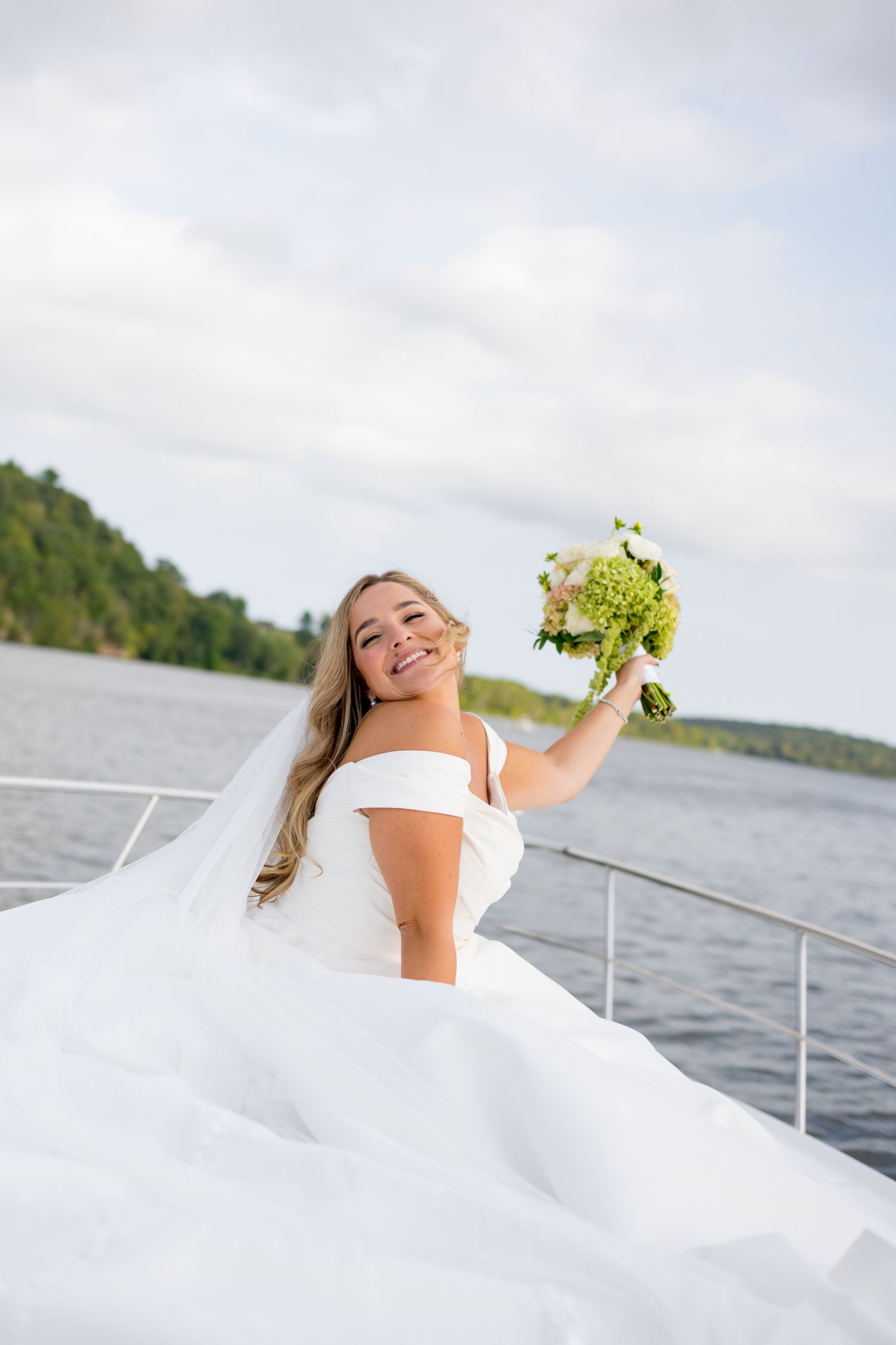 Bride in flowing white dress celebrates joyfully on yacht deck with bouquet.