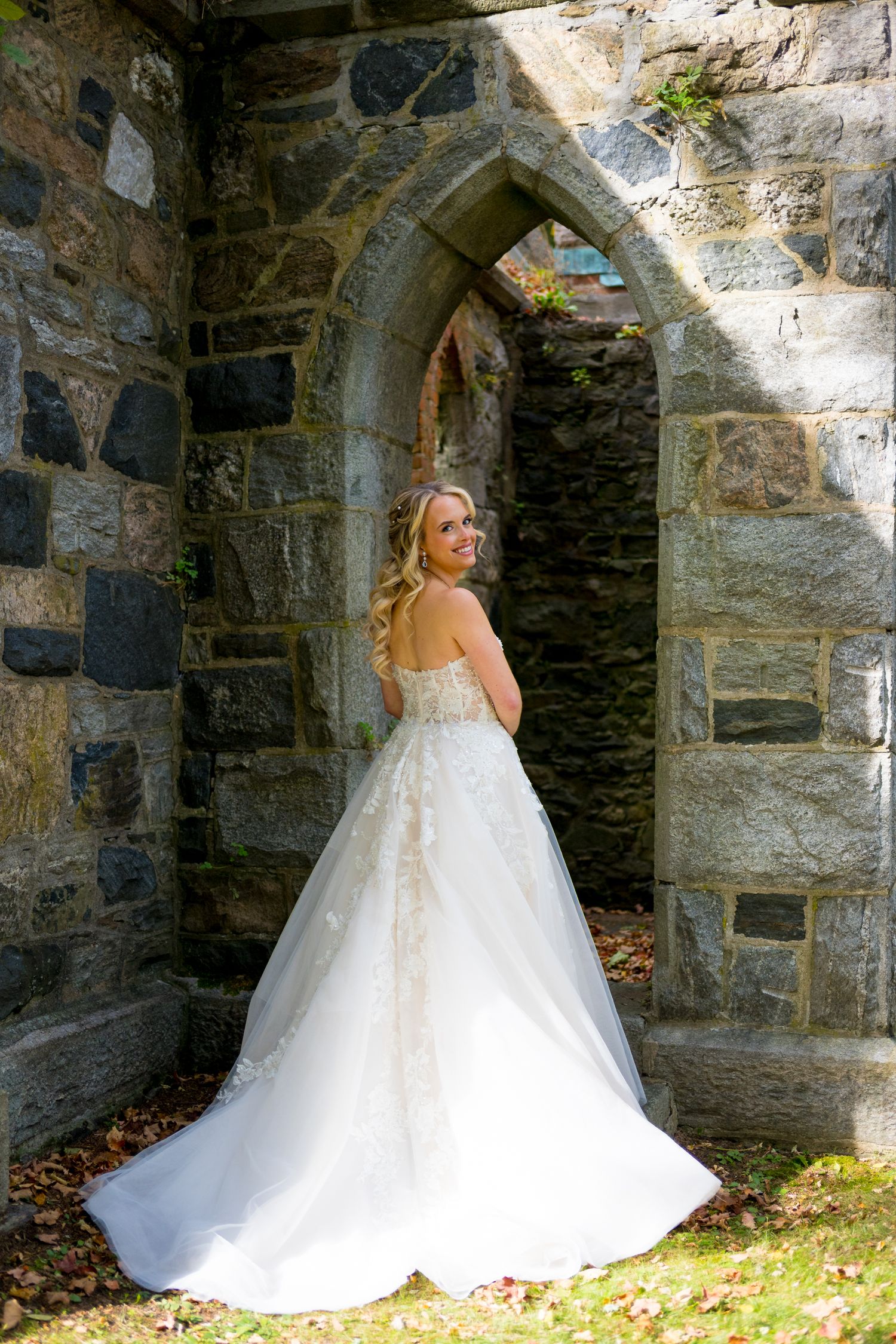A bride in a flowing white wedding gown stands beneath a stone archway at a historic church or castle.