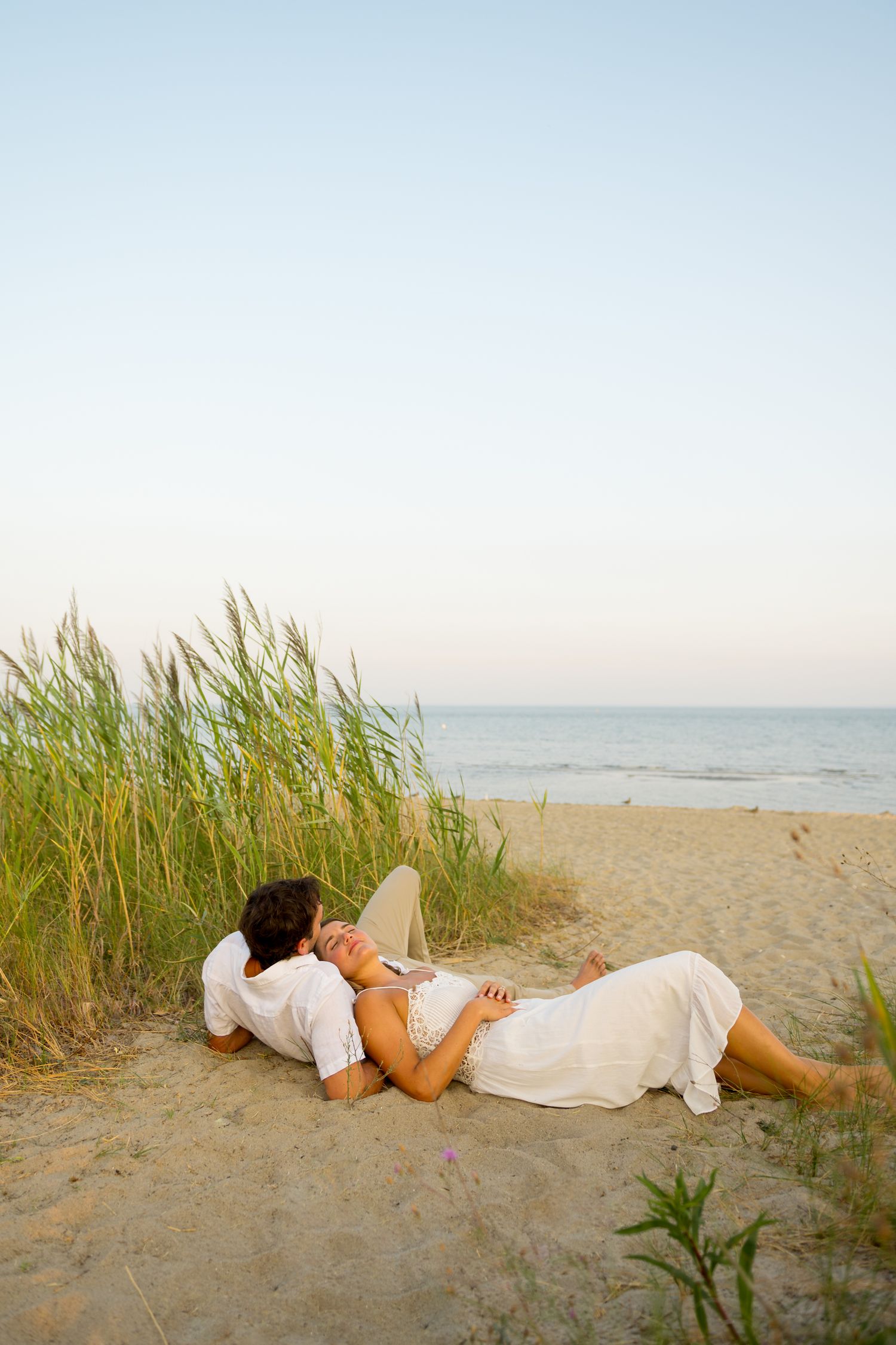 Couple in white clothing lying on sandy beach near tall grass at sunset.