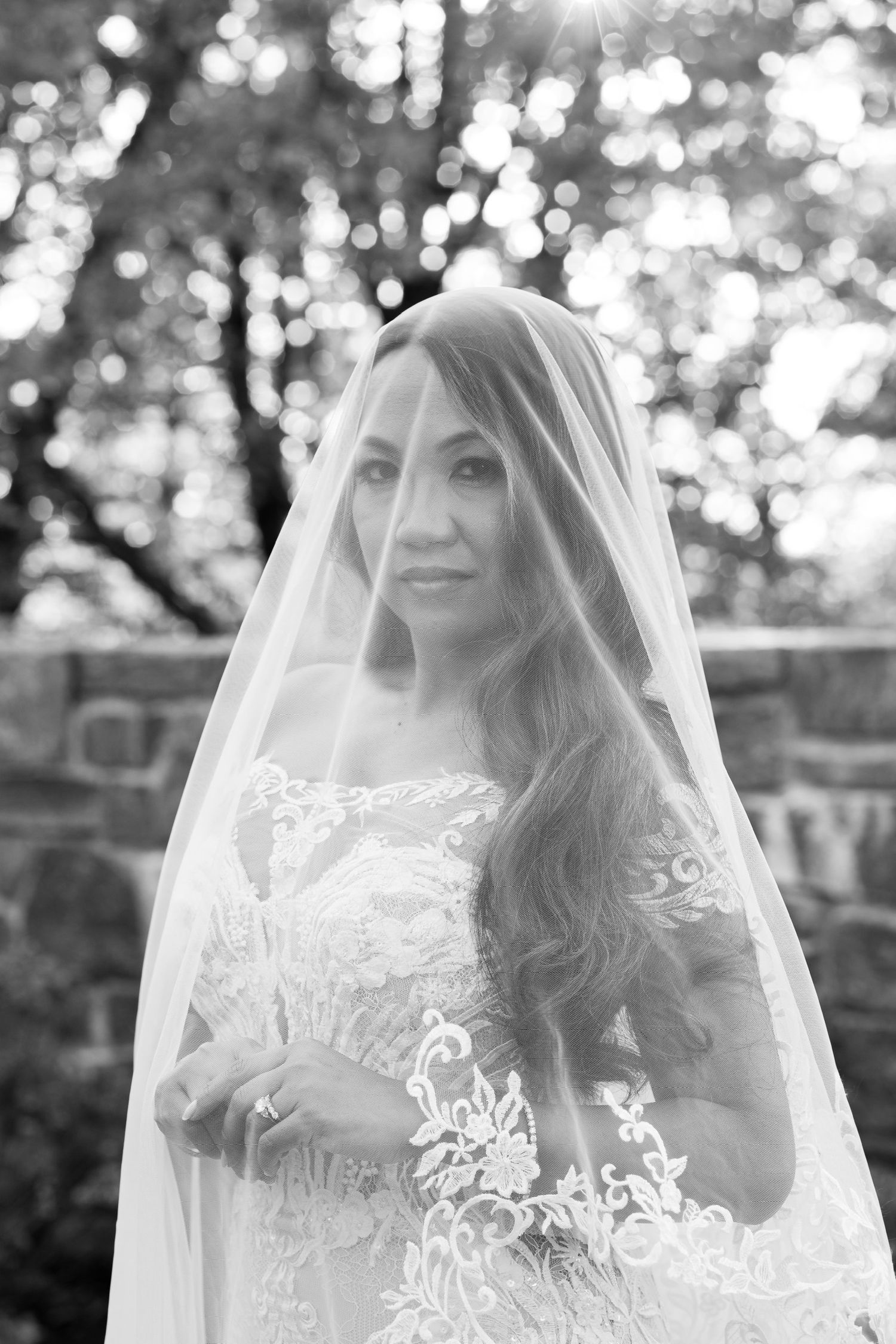 A bride wearing an elegant lace gown and veil poses in front of a stone wall at Le Chateau in Westchester County, New York.