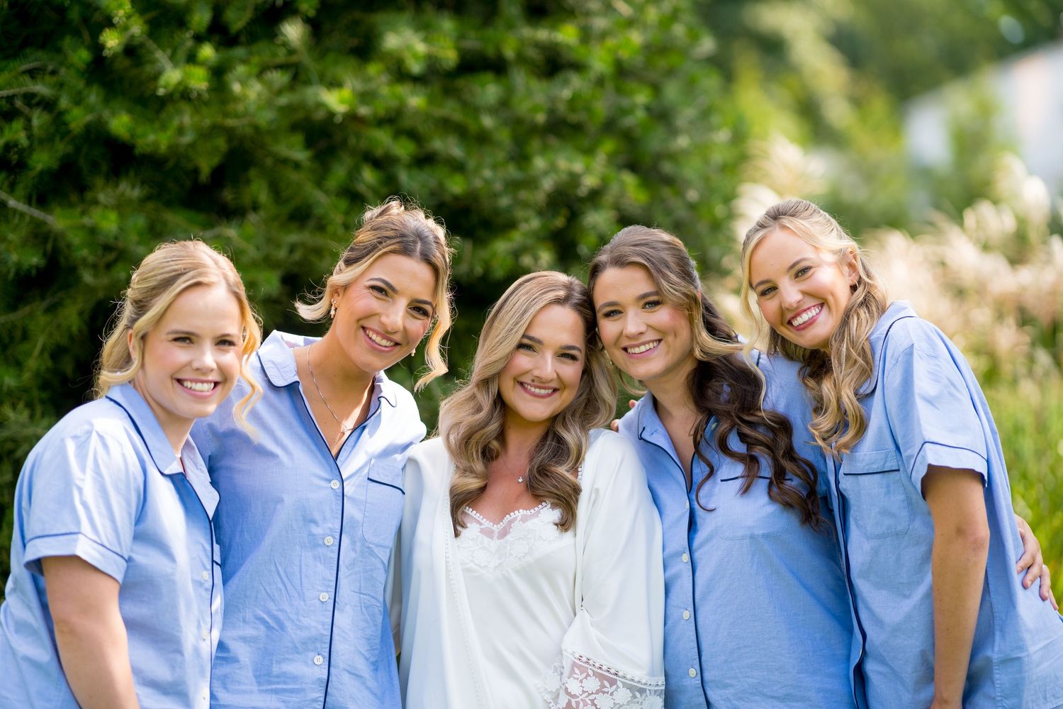 Group wearing light blue tops pose together outdoors with greenery in background.