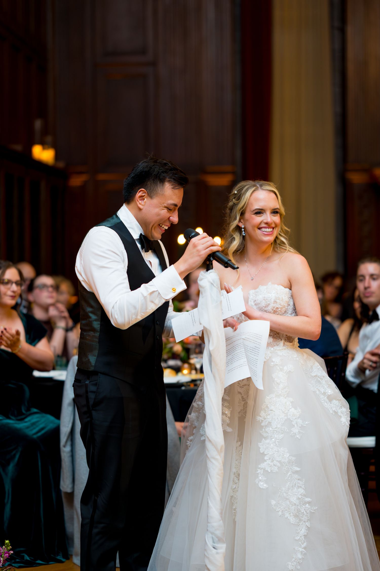 A joyful wedding reception moment as newlyweds share a laugh while speaking into microphones during their celebration.
