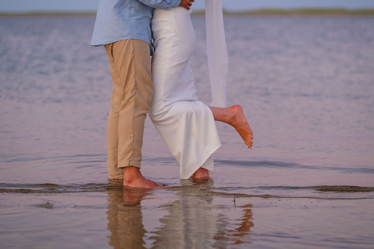People's feet standing in shallow ocean water at sunset with reflections on the wet sand.