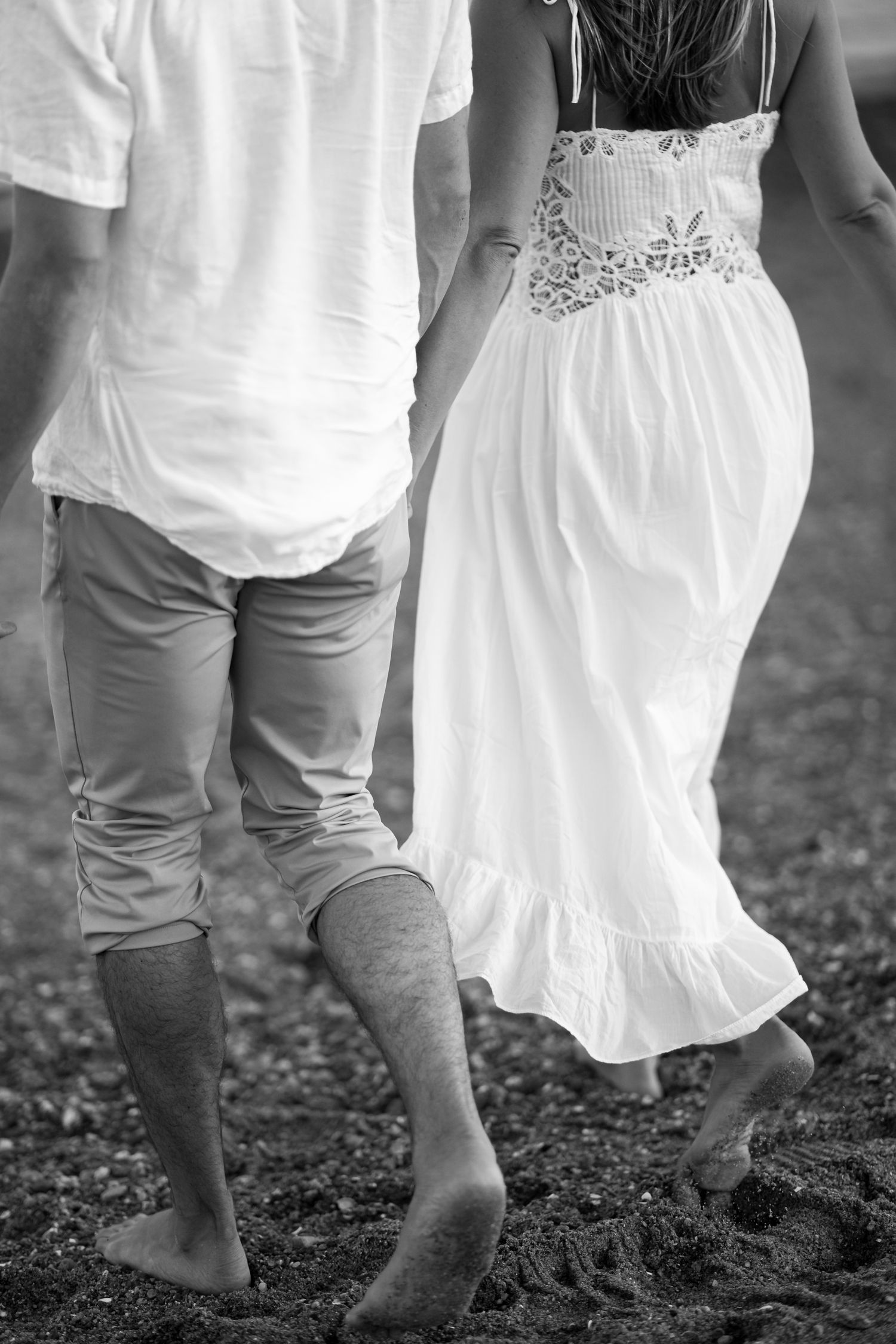 Romantic couple walking barefoot on beach with white dress and casual attire in black and white.