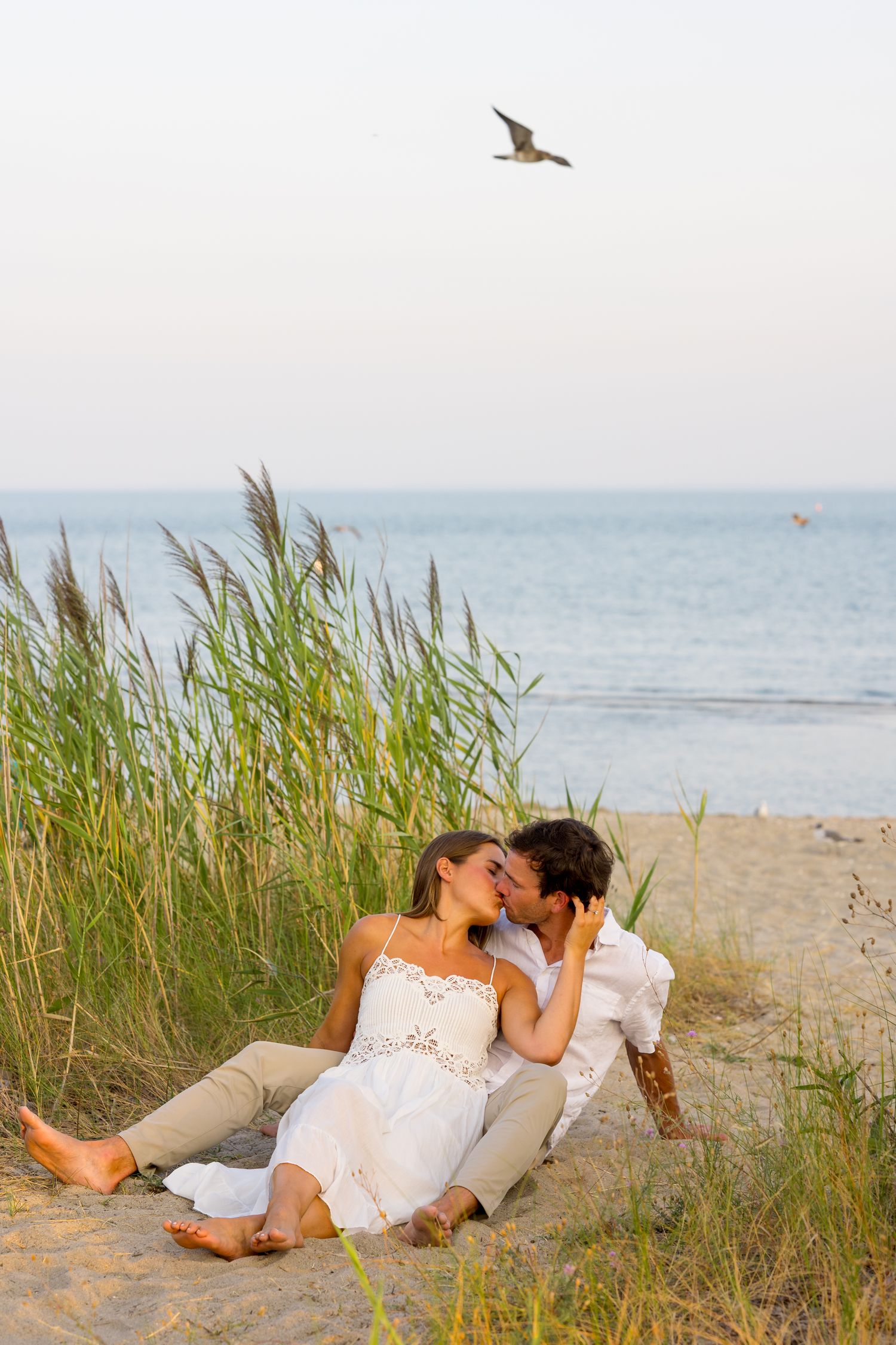 A romantic couple lounges together in white outfits among beach grass at sunset by the ocean.