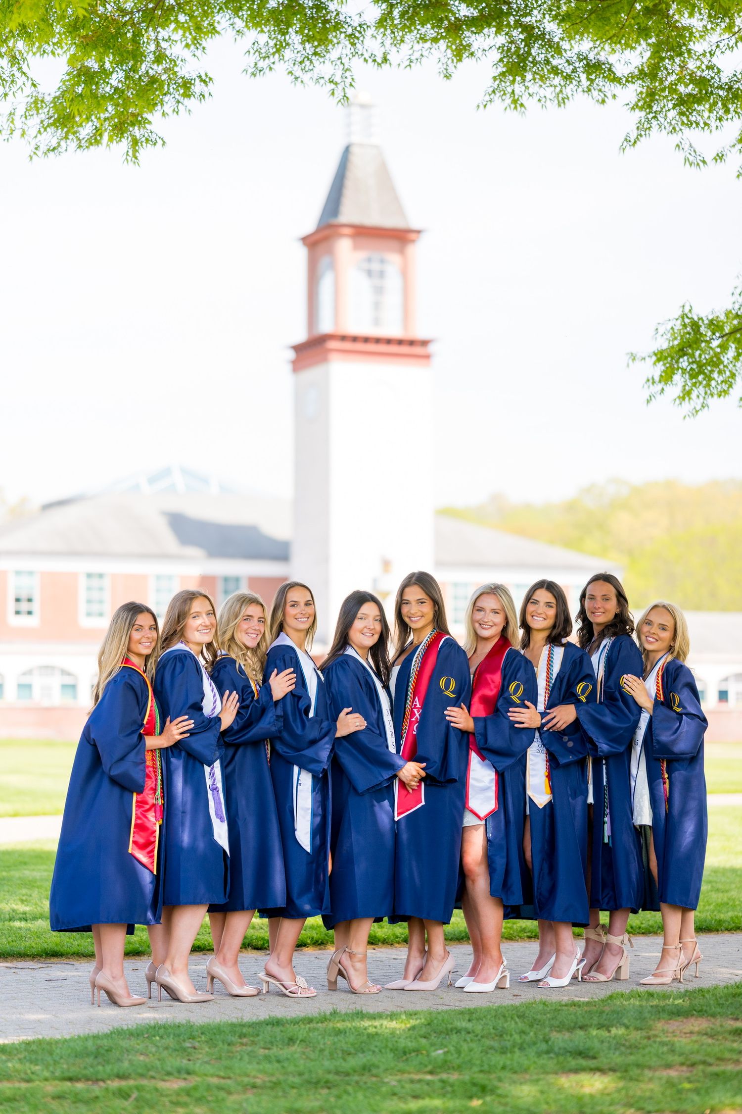 Quinnipiac University graduates in blue caps and gowns pose together on campus lawn with church steeple in background.