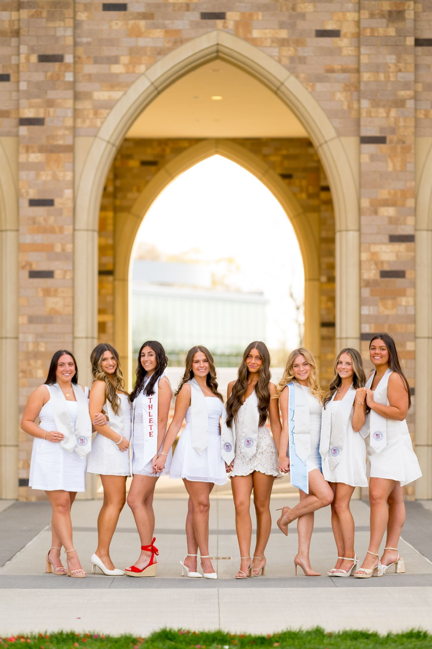 Sacred Heart University graduates in white dresses pose together under stone archways for graduation photos.