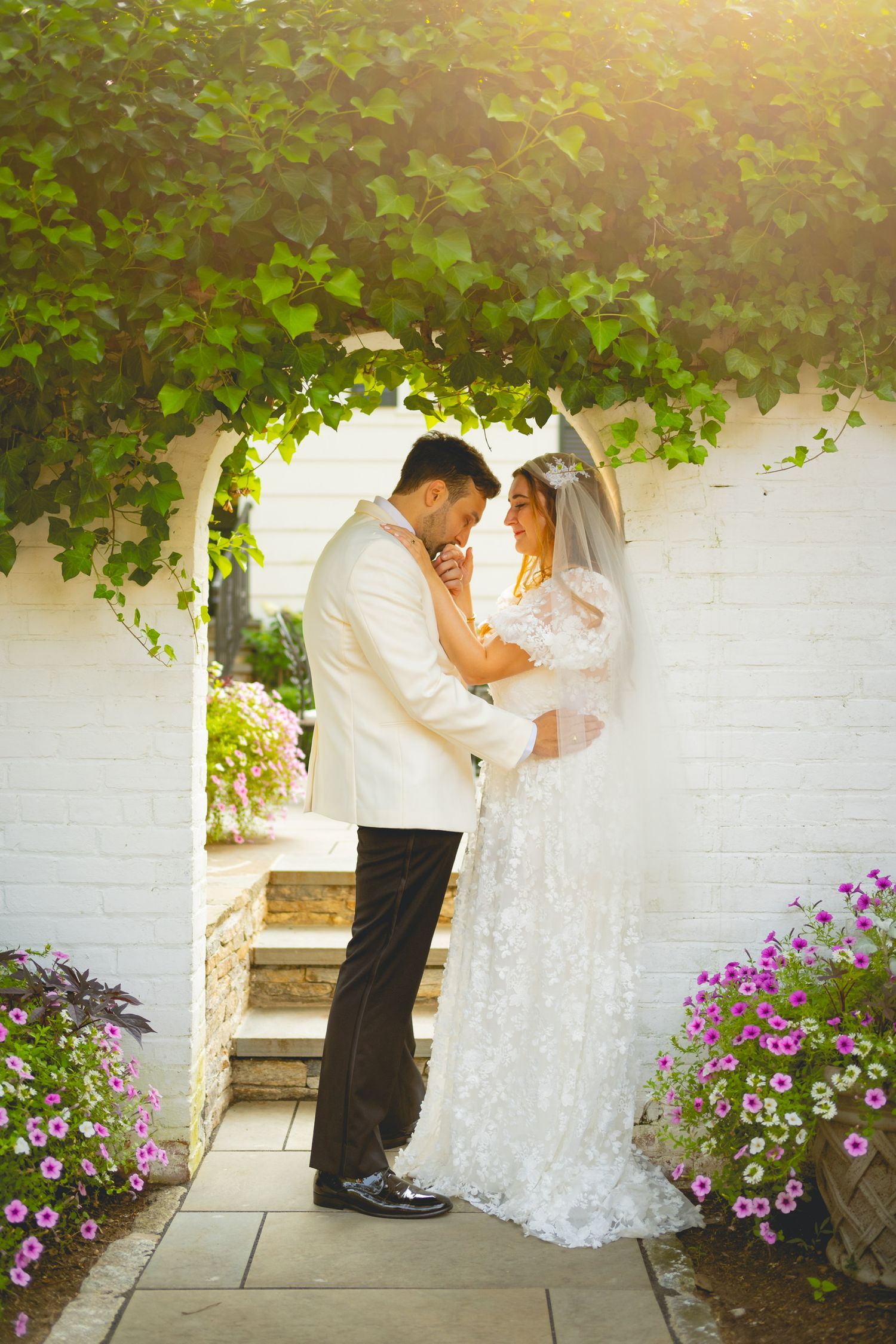 Romantic wedding moment against a white wall with purple flowers and green vines.