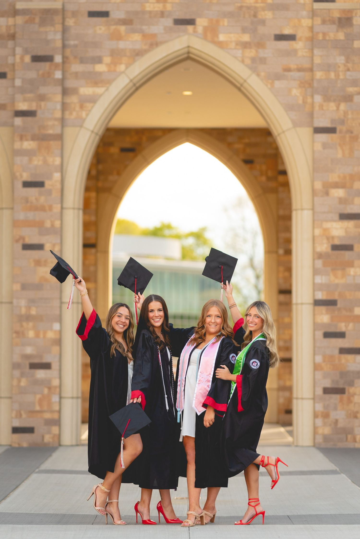 Graduates in black caps and gowns with red heels pose playfully under stone archways.