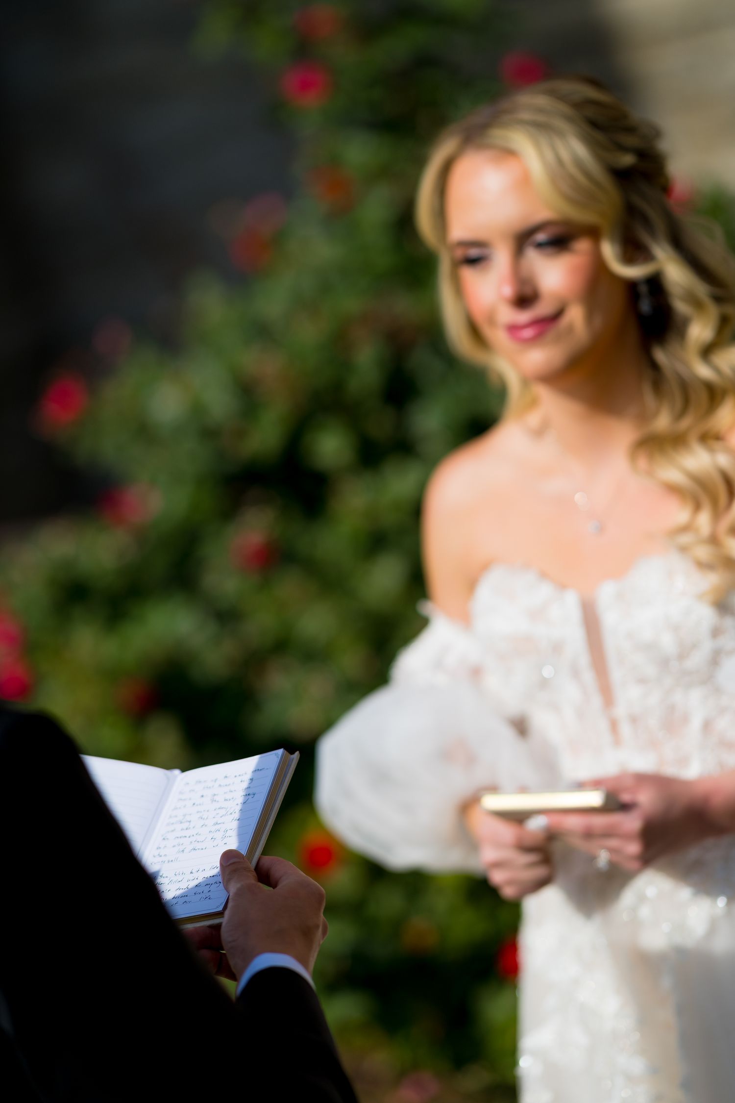 A bride in a white lace dress reads from a small book in a garden setting with red flowers in the background.