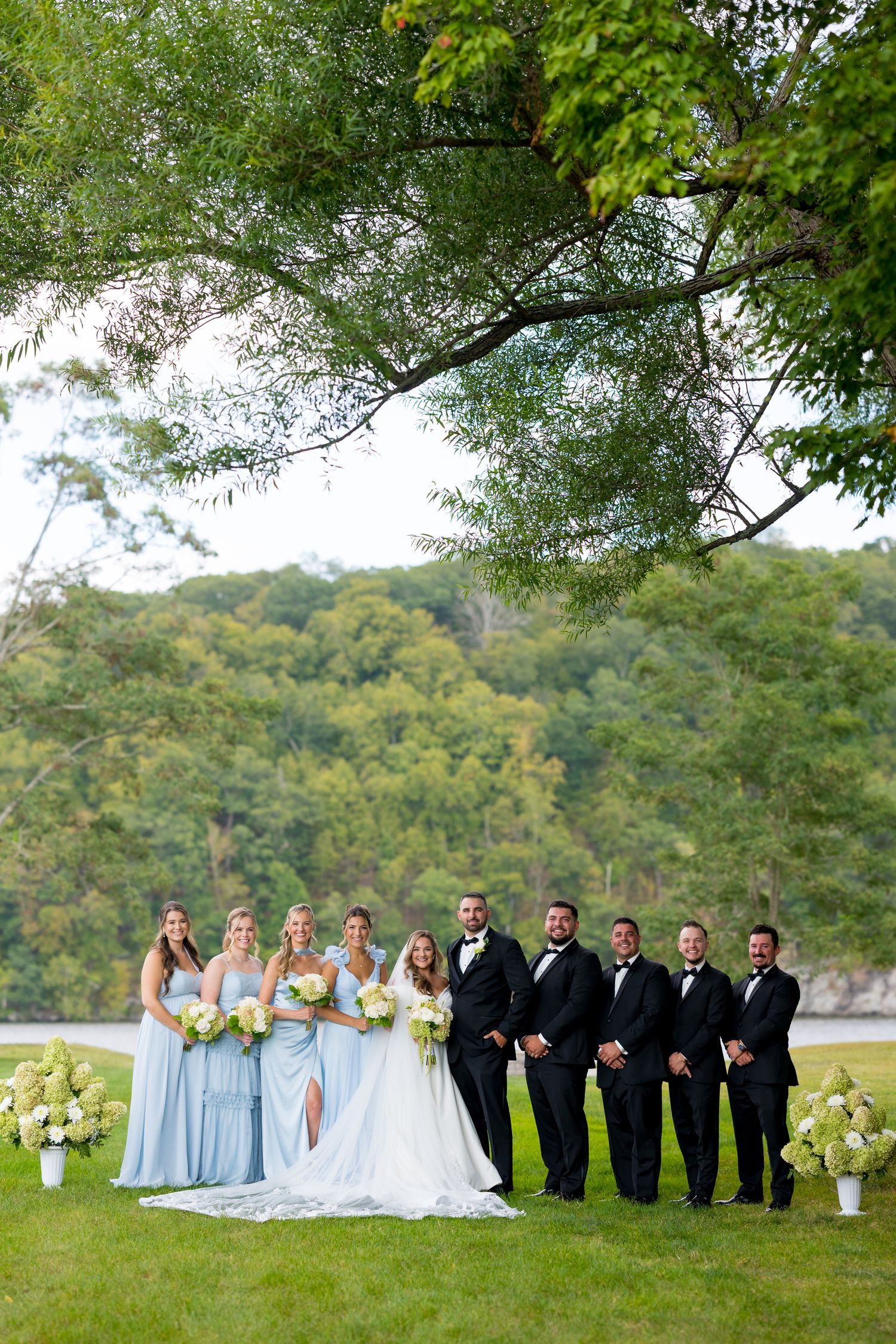 Wedding party in light blue bridesmaid dresses and black tuxedos pose outdoors with mountain backdrop.