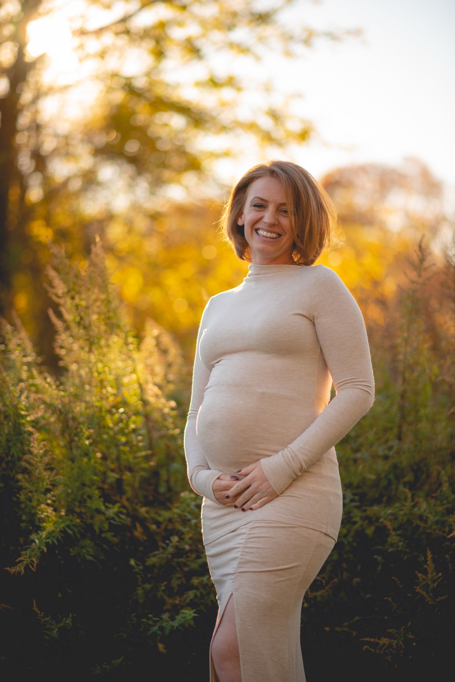 Expectant mother in cream sweater poses joyfully during sunset maternity photoshoot.