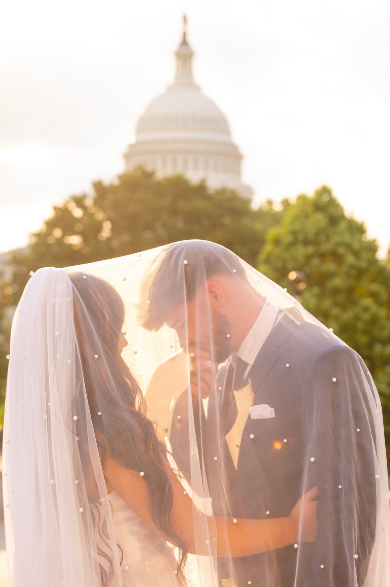 Wedding couple silhouetted under veils with sunset and domed building in background.