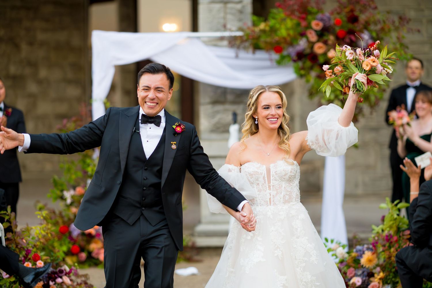 A joyful wedding couple celebrates with guests during an outdoor ceremony surrounded by colorful flowers and stone walls.