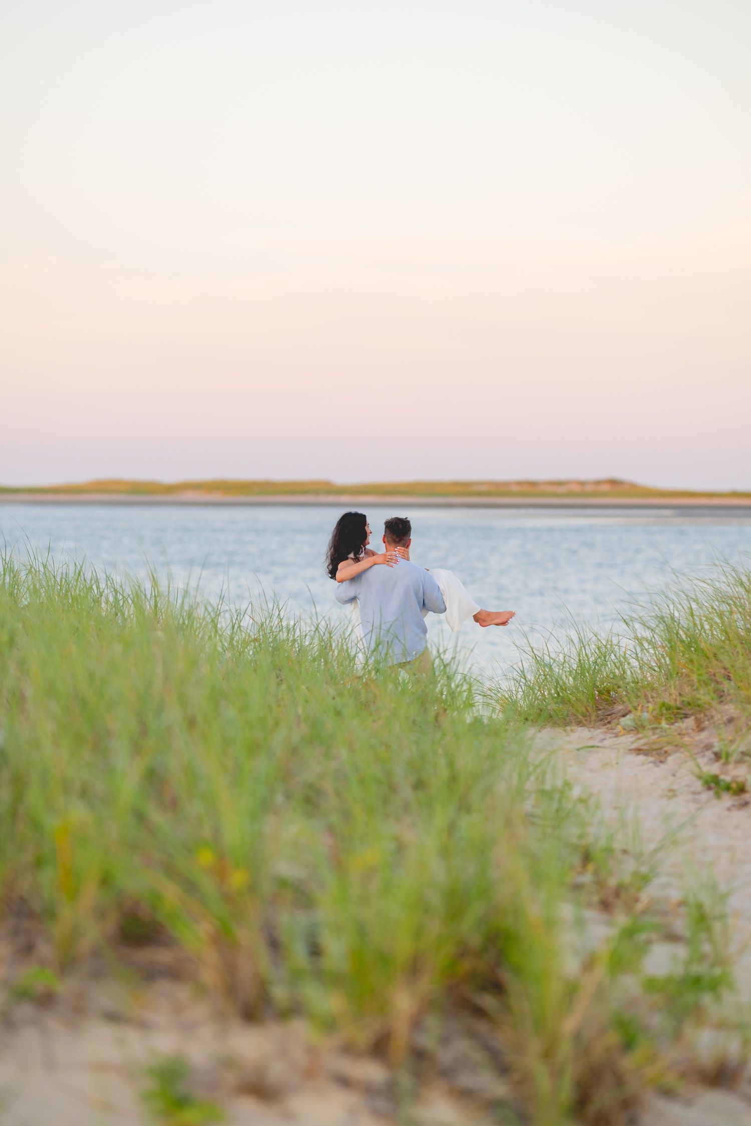 Two people sit together on a beach at sunset with tall grass in the foreground and calm waters in the distance.