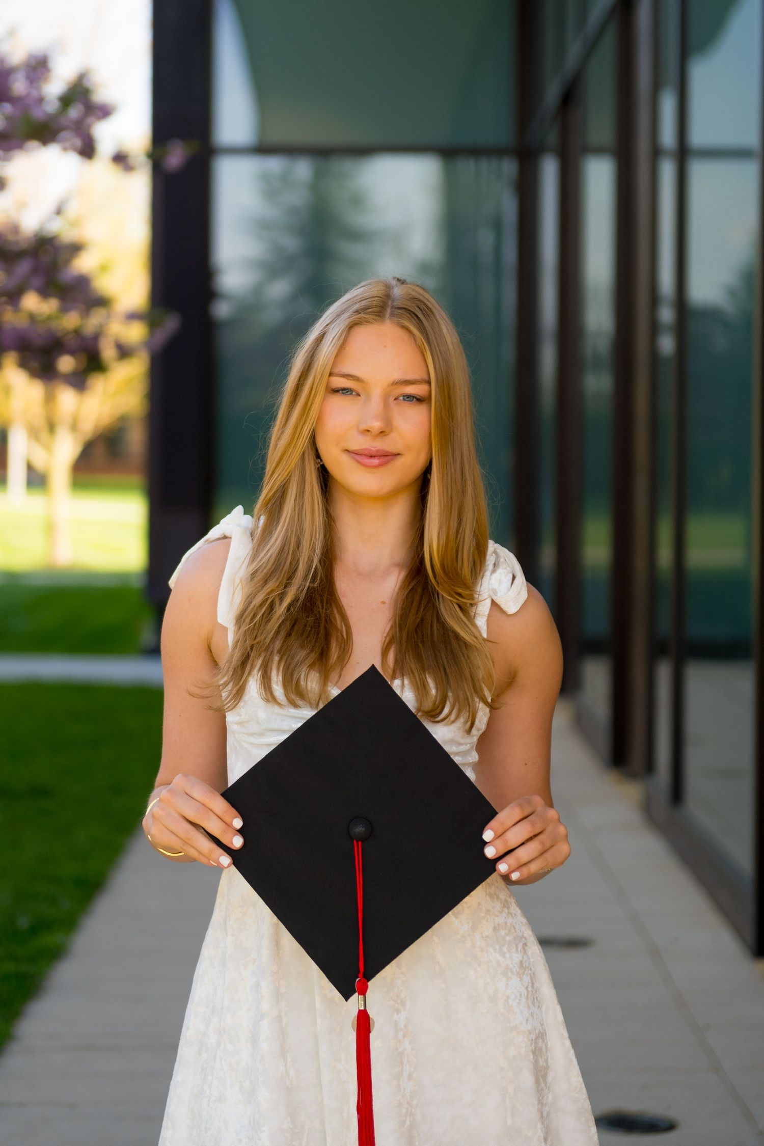 Sacred Heart University graduate in white dress poses with black graduation cap during outdoor photo session.