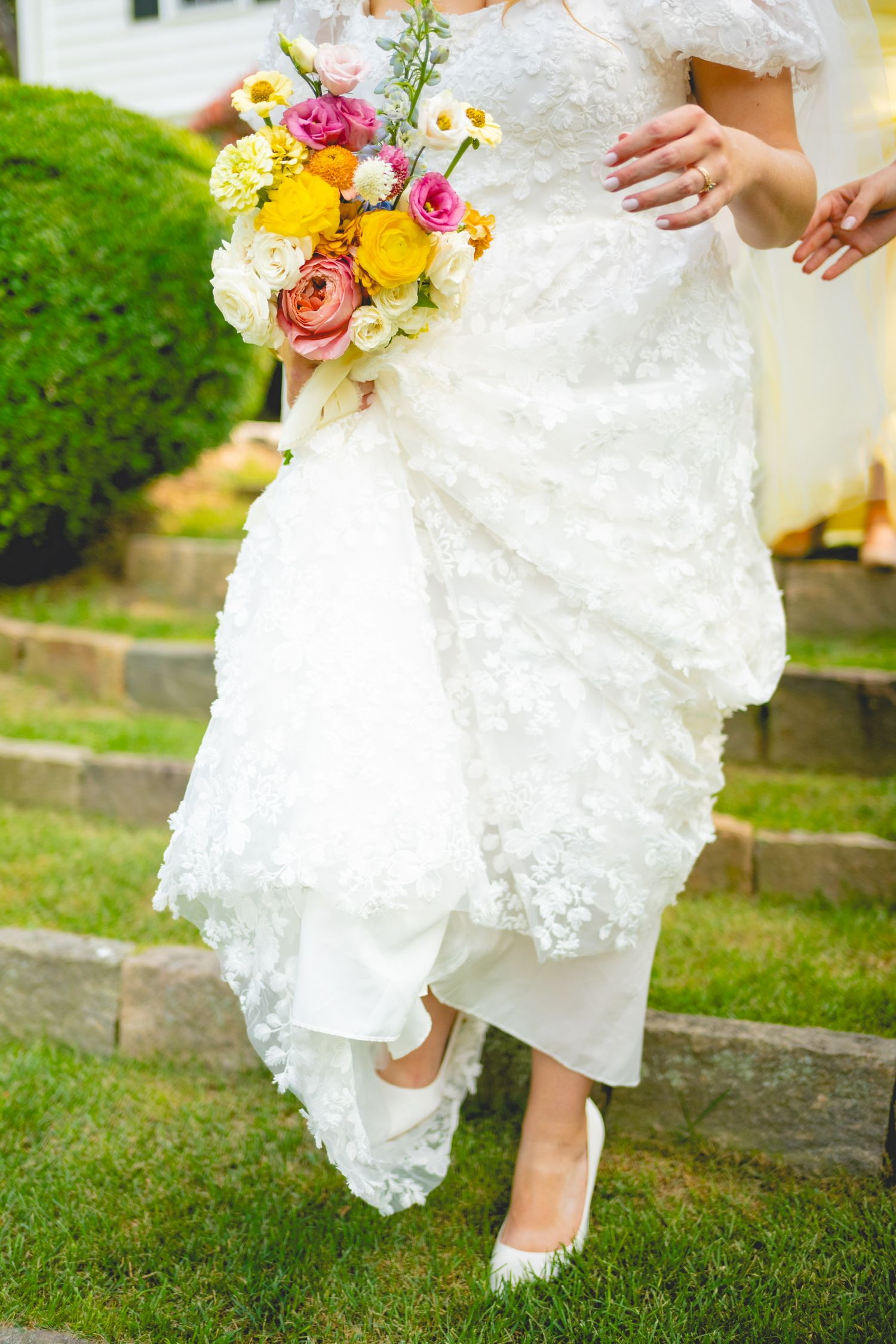 White lace wedding dress and colorful bouquet details against green grass.