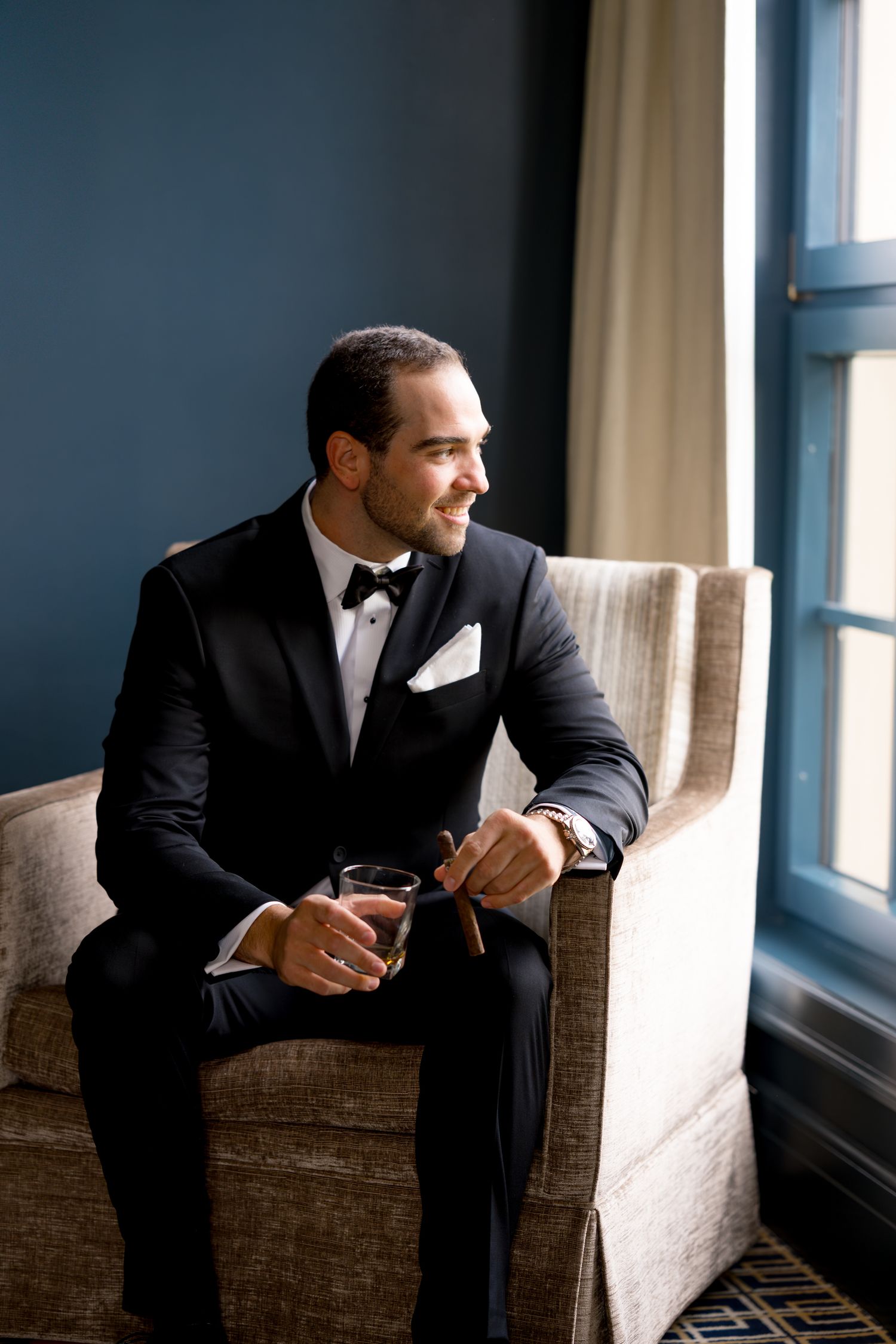 Groom in formal attire sits thoughtfully by window in moody lighting.