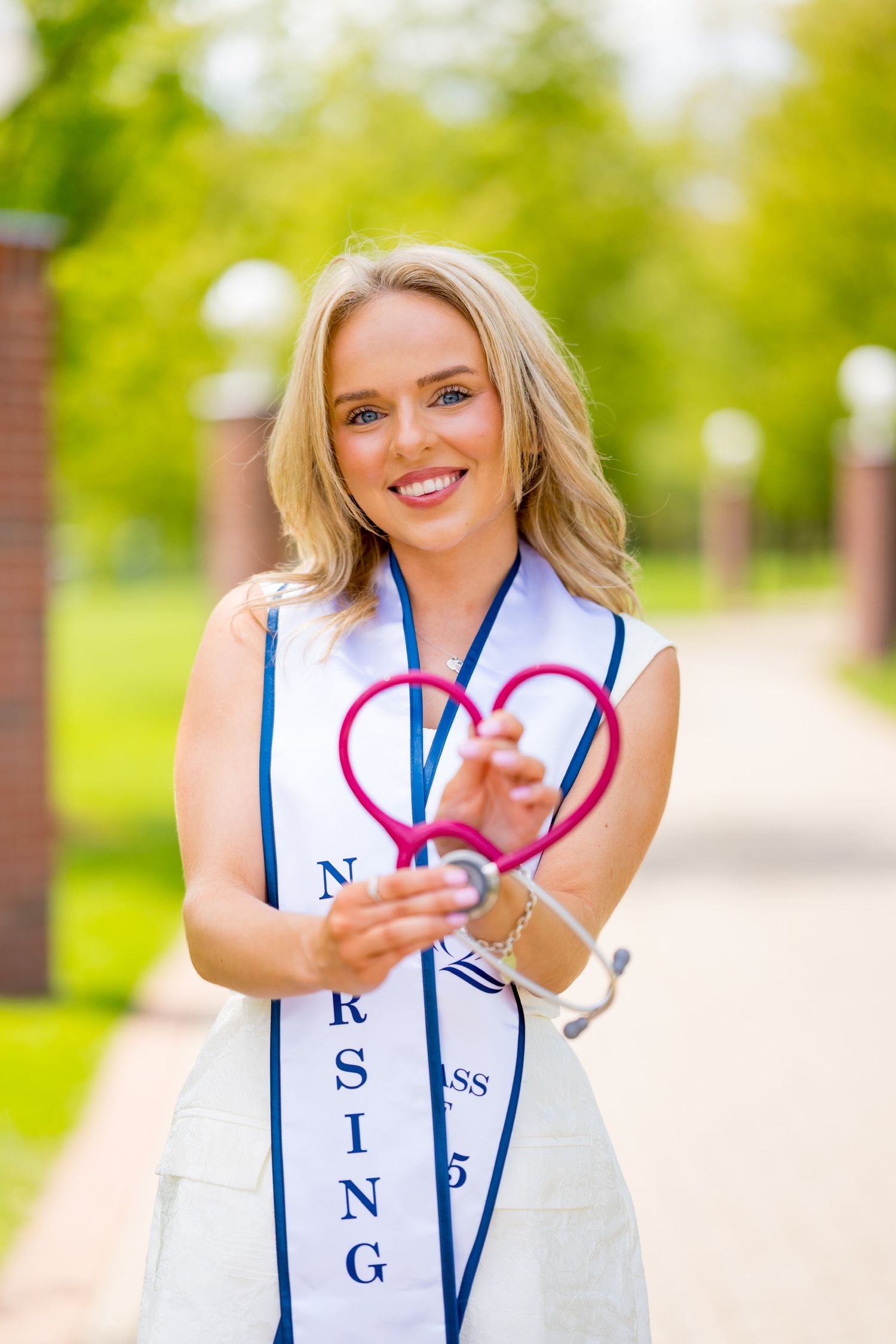 Quinnipiac University graduate in white dress and blue sash making heart sign with stethoscope outdoors on sunny day.