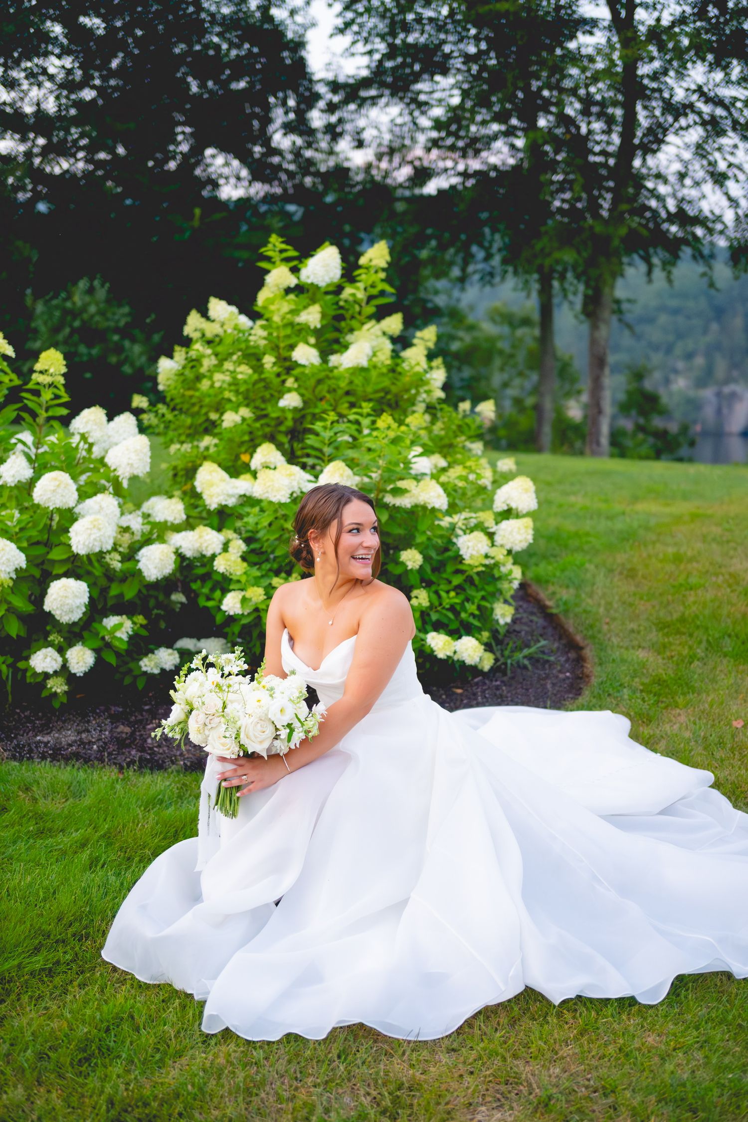 Bride in white gown lying on grass surrounded by white hydrangea bushes in garden setting.