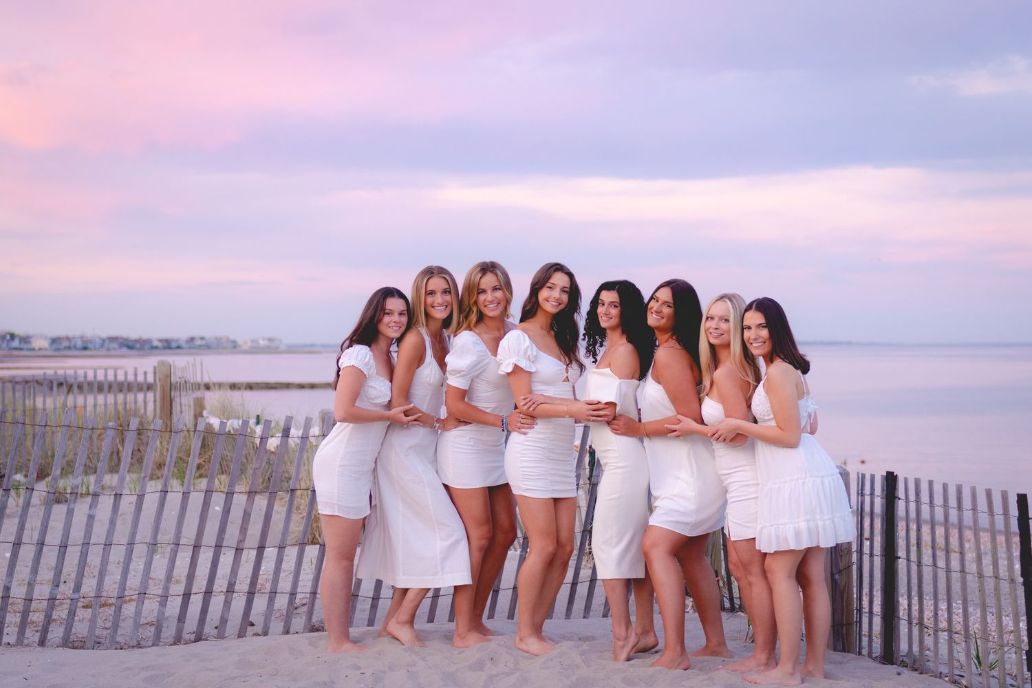 Fairfield University graduates in white dresses posing on a beach at sunset.