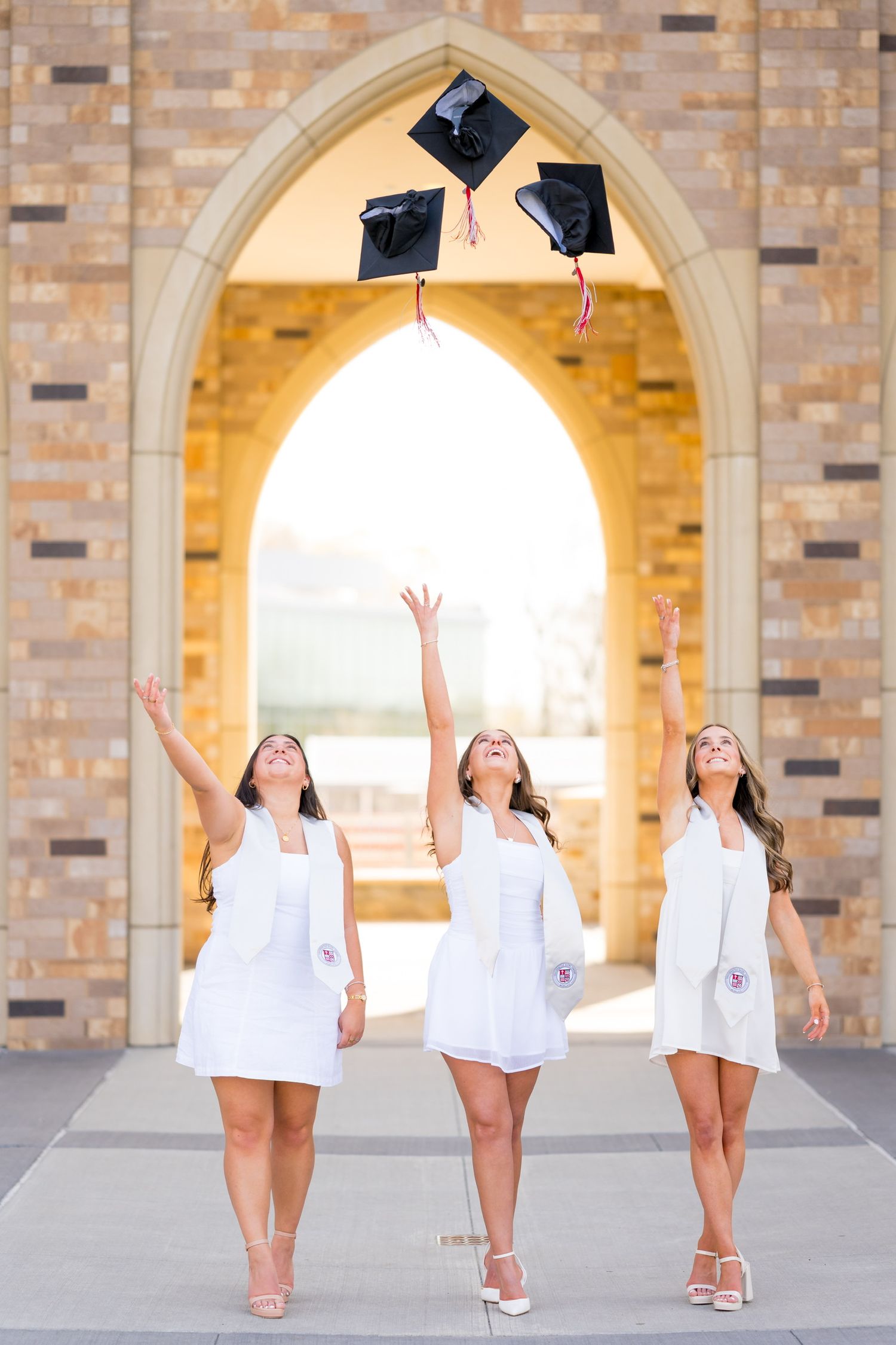 Sacred Heart graduates throwing caps in celebration under stone archways for graduation photos.