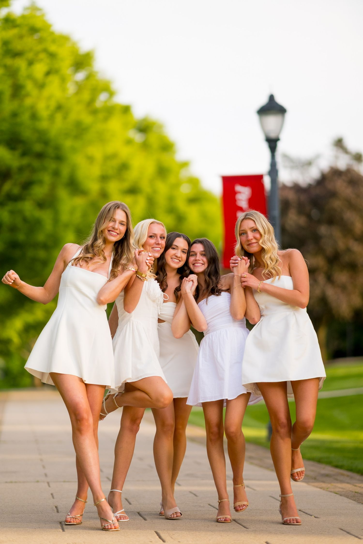 Fairfield University graduates in white dresses poses playfully on campus sidewalk near lamppost.