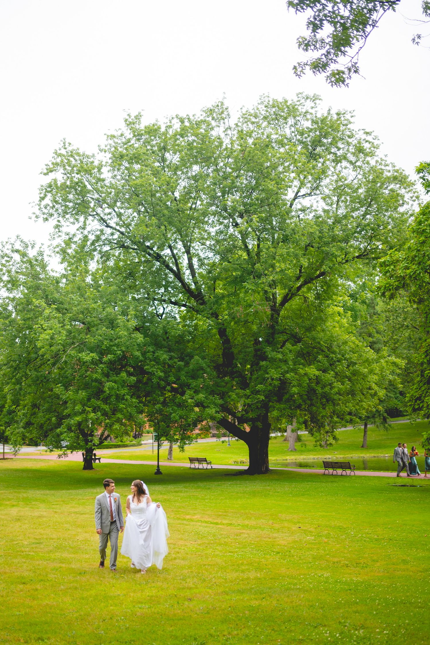 Wedding couple walking across green lawn with trees in background.