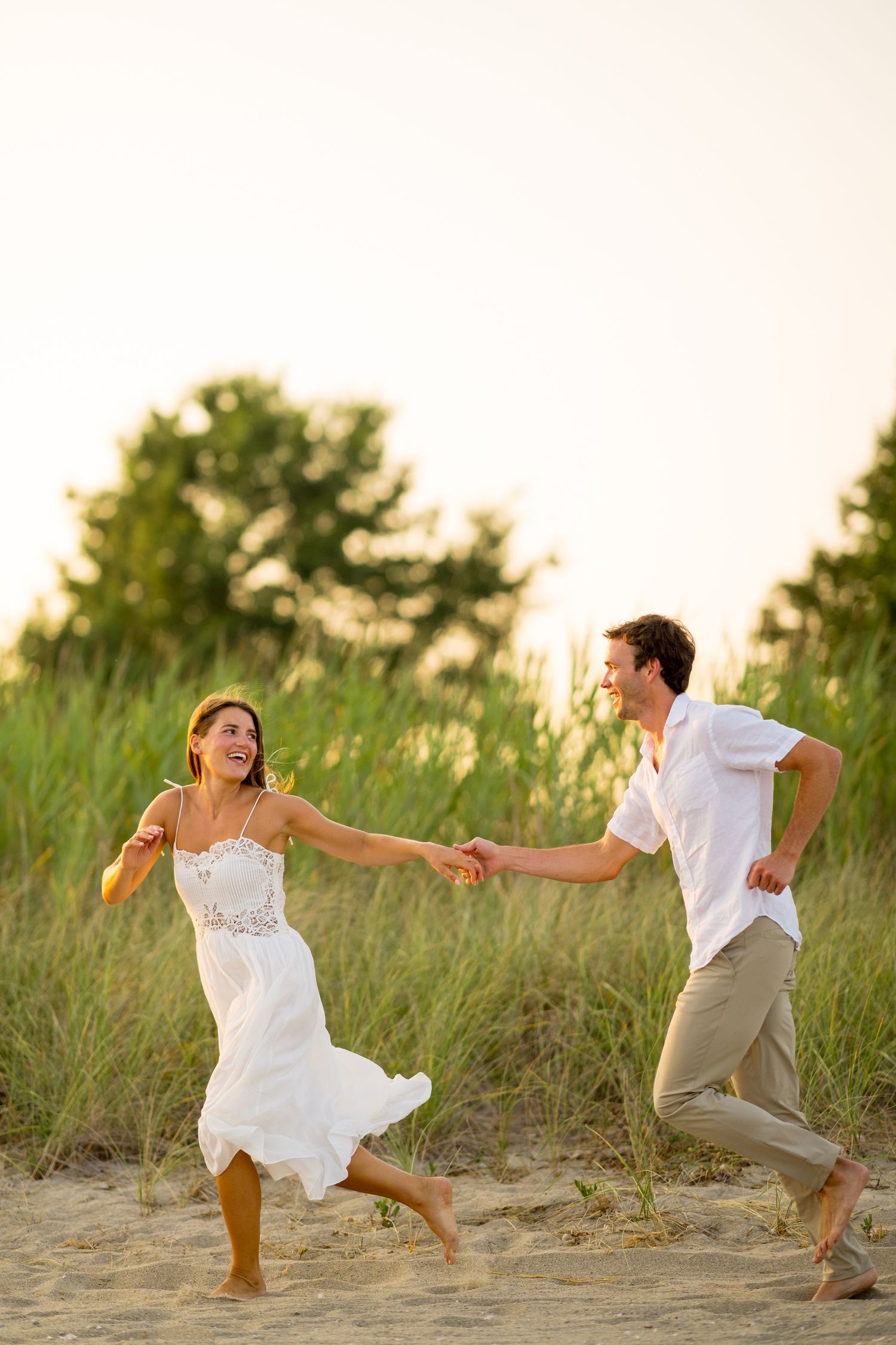 A couple in white and beige outfits dances playfully on sand dunes with beach grass in the background.