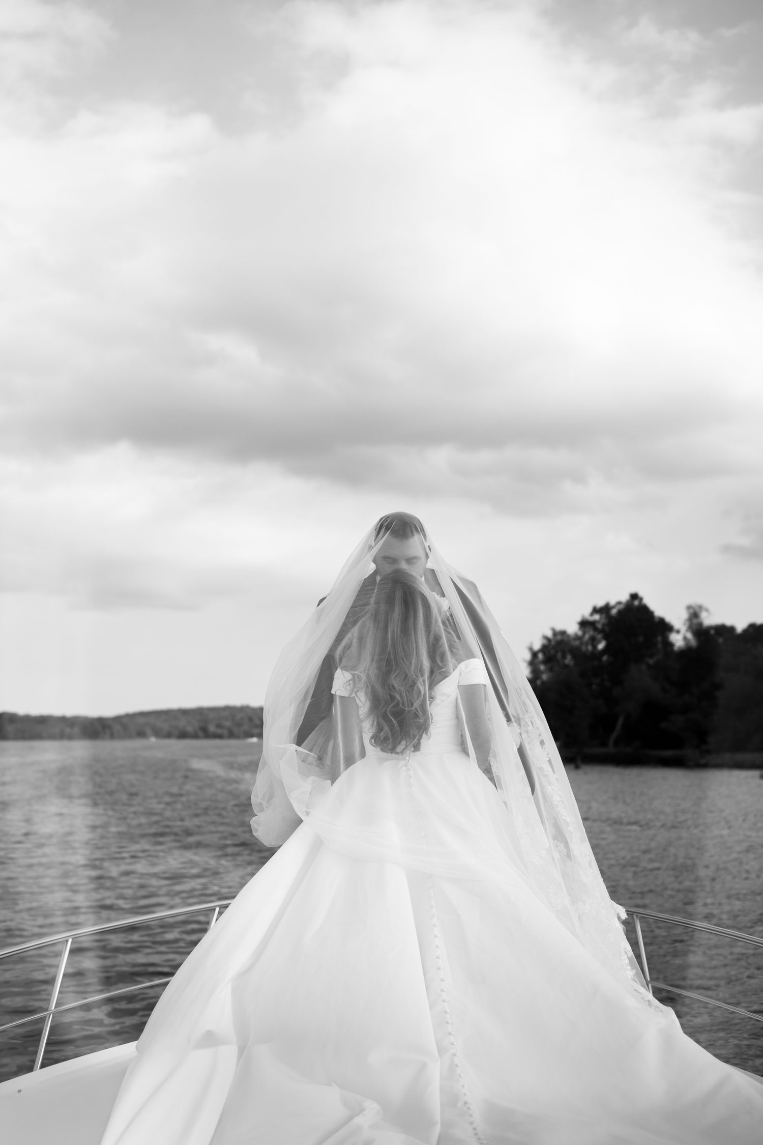 A bride in a flowing white dress poses on a boat with water and clouds in background.